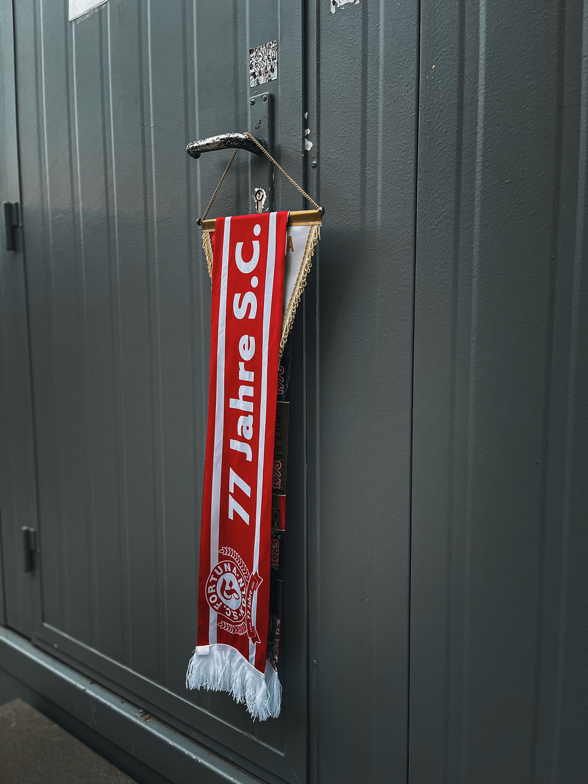 Rot-weißer Jubiläumsschal mit der Aufschrift „77 Jahre S.C.“, über einem Wimpel mit goldener Kette und weißen Fransen hängend.