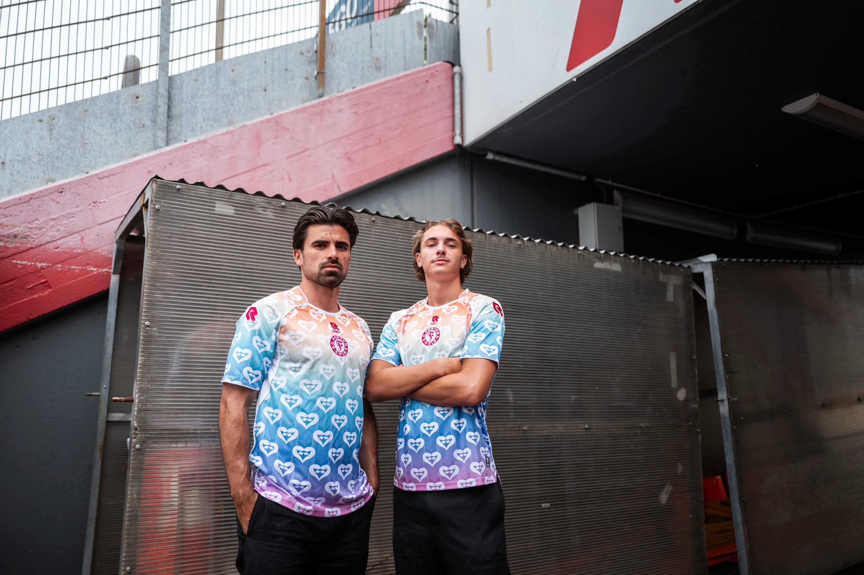 Zwei SC Fortuna Köln Spieler im Pride-Trikot stehen nebeneinander im Außenbereich des Stadions vor einer Metallwand.
