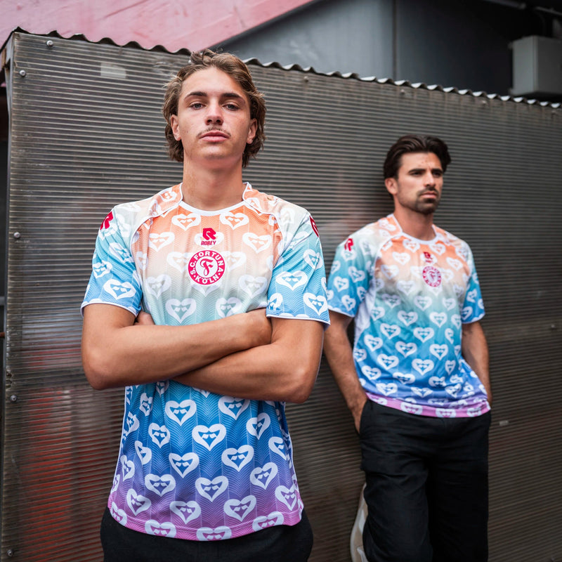 Zwei Spieler stehen nebeneinander im Pride Shirt von Fortuna Köln mit Herzmuster und mehrfarbigem
Farbverlauf. Das rote Vereinslogo ist
zentral auf der Brust platziert.