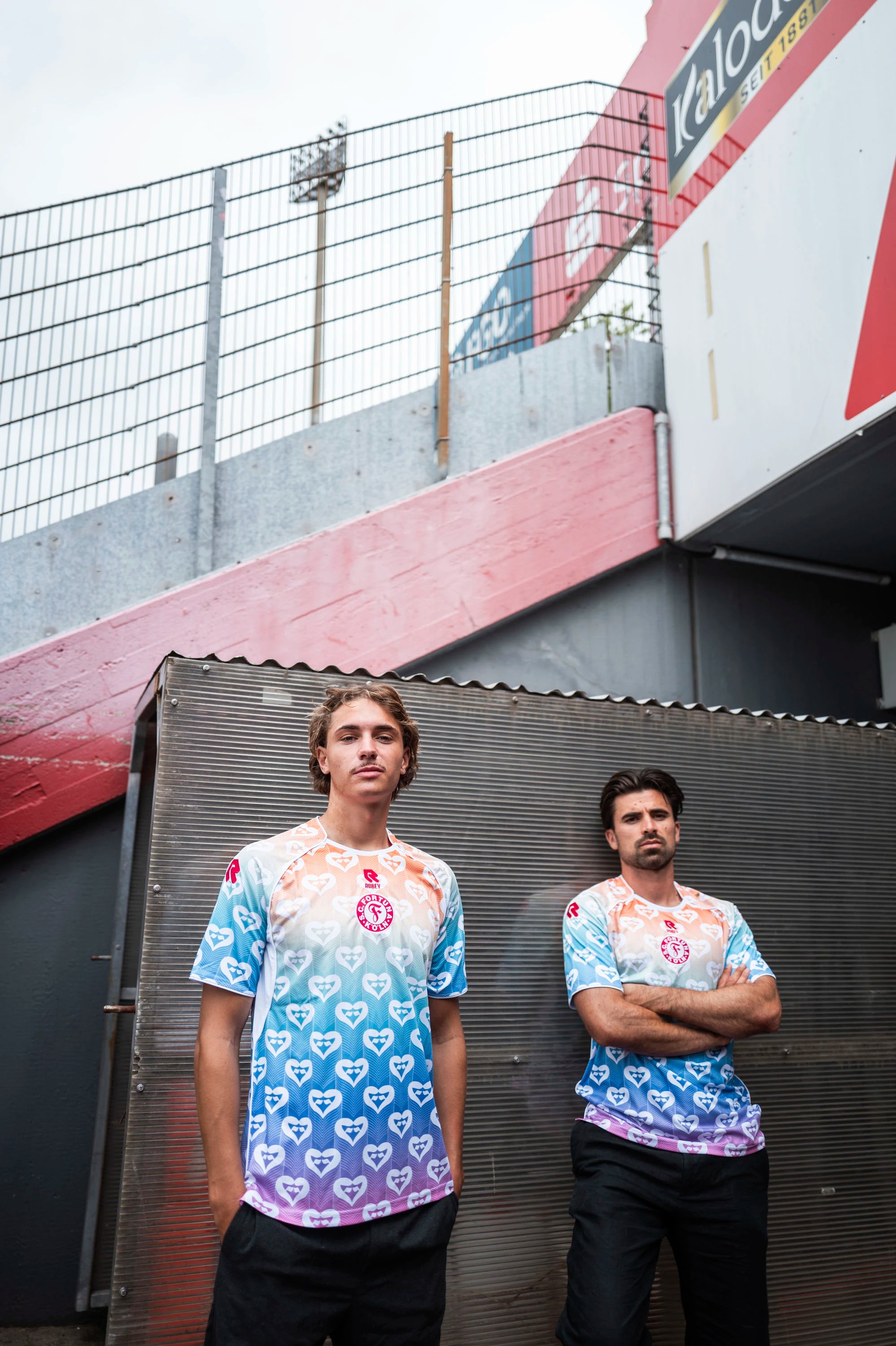 Zwei Spieler des SC Fortuna Köln posieren im Stadionbereich im Pride-Trikot, im Hintergrund sind Stadiontreppe und Werbebanden zu sehen.