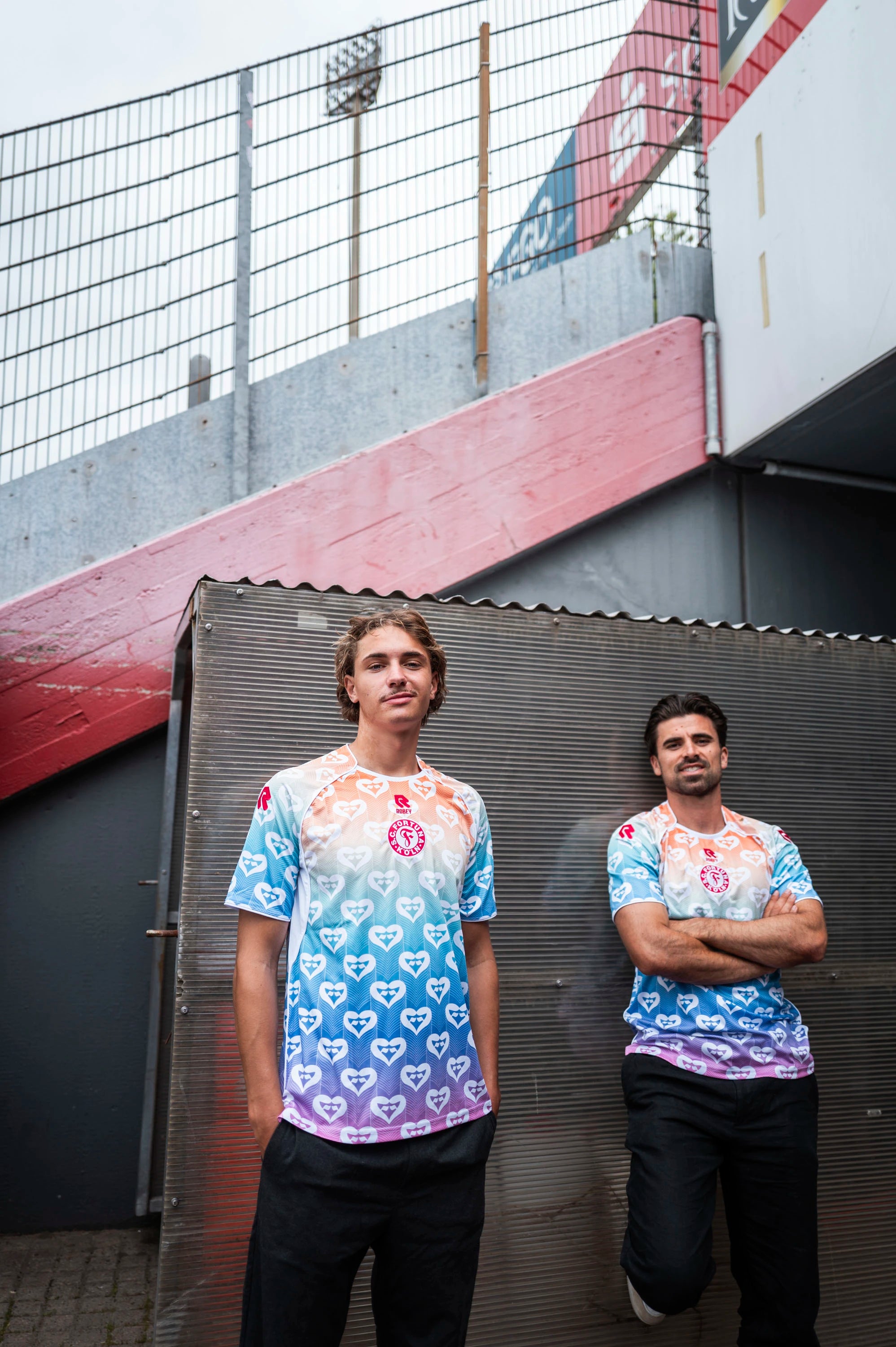 Zwei Spieler tragen das Pride Shirt
von Fortuna Köln mit Farbverlauf von
Weiß über Rot und Blau zu Lila. Das
Muster besteht aus wiederholten
Herzsymbolen, auf der Brust befindet
sich das rote Fortuna-Logo.