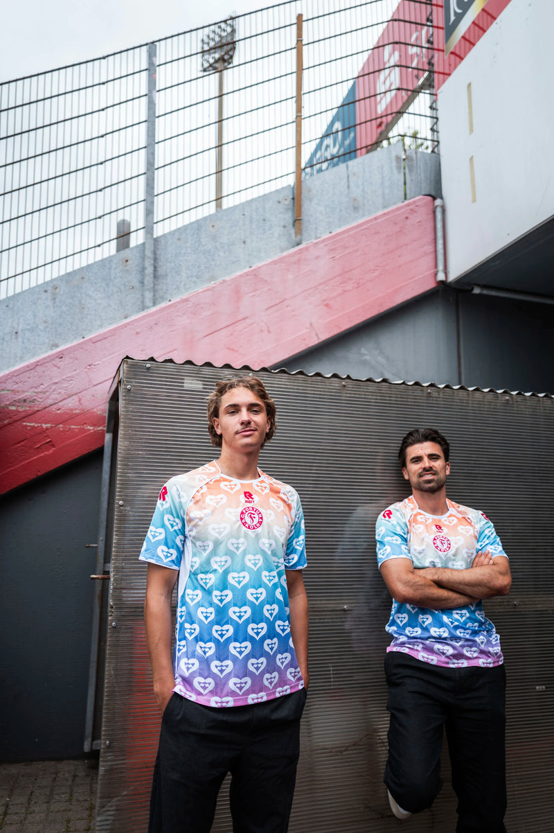 Zwei Spieler tragen das Pride Shirt
von Fortuna Köln mit Farbverlauf von
Weiß über Rot und Blau zu Lila. Das
Muster besteht aus wiederholten
Herzsymbolen, auf der Brust befindet
sich das rote Fortuna-Logo.