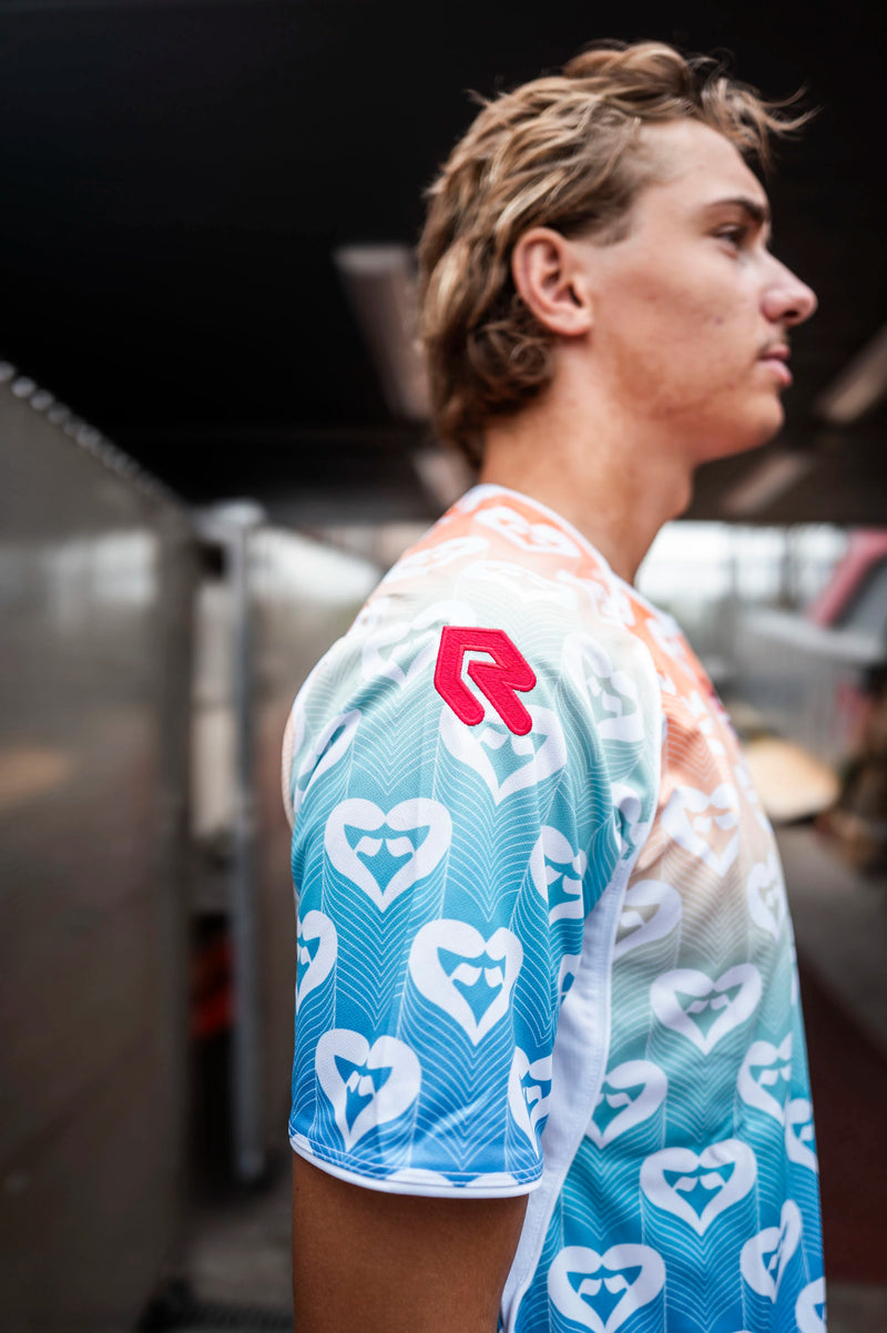 Seitenansicht eines SC Fortuna Köln Spielers im Pride-Trikot. Deutlich erkennbar sind das rote Robey-Logo auf dem Ärmel, der Farbverlauf von Orange zu Blau und das weiße Herzmuster.