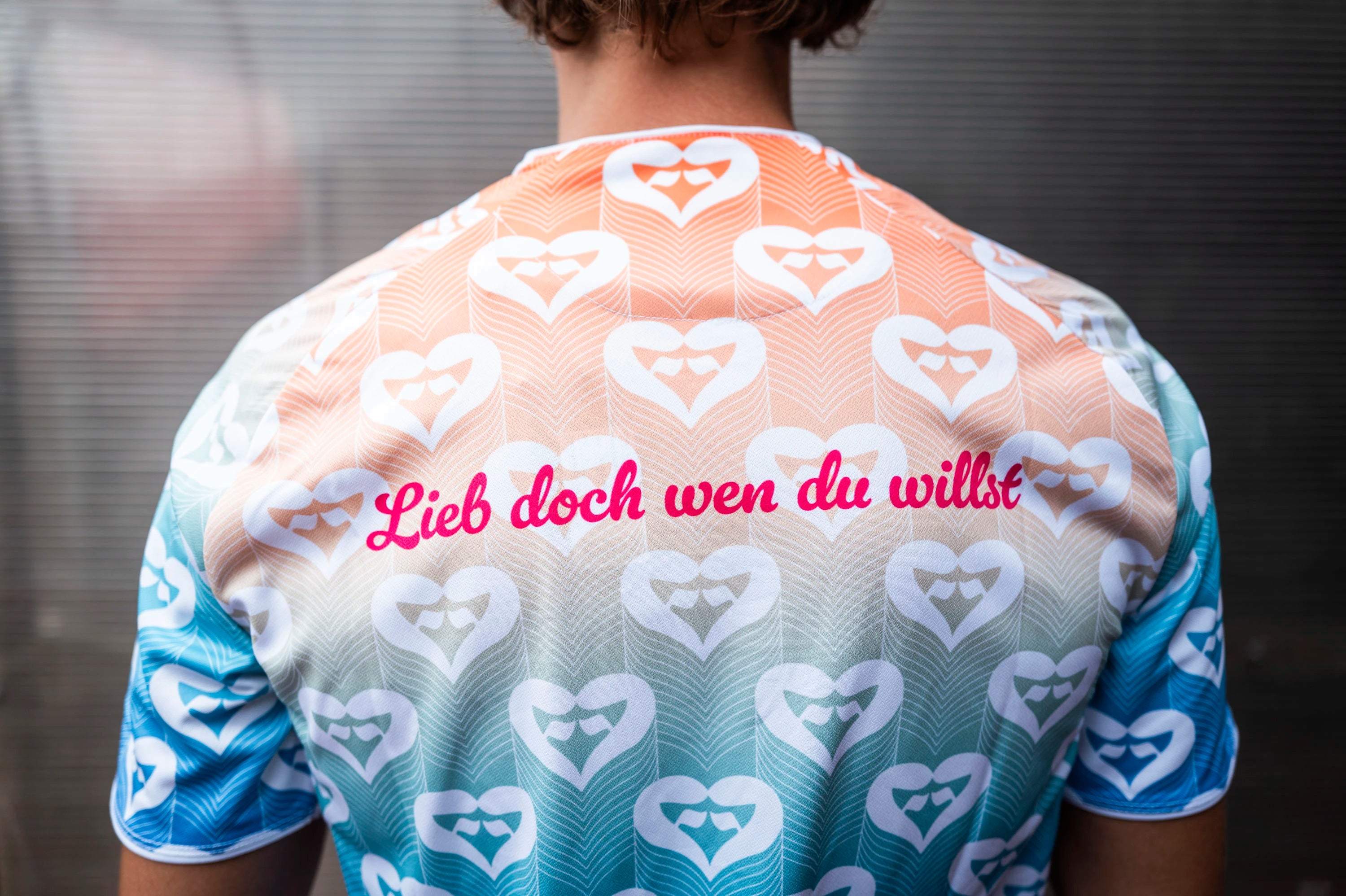 Rückansicht des Pride-Trikots,
getragen von einem Spieler des SC
Fortuna Köln. Auf dem oberen Rücken
steht in pinker Schrift der Slogan „Lieb Pride Shirt – SC Fortuna Köln Fanshop
doch wen du willst“ über dem durchgehenden Herzmuster.