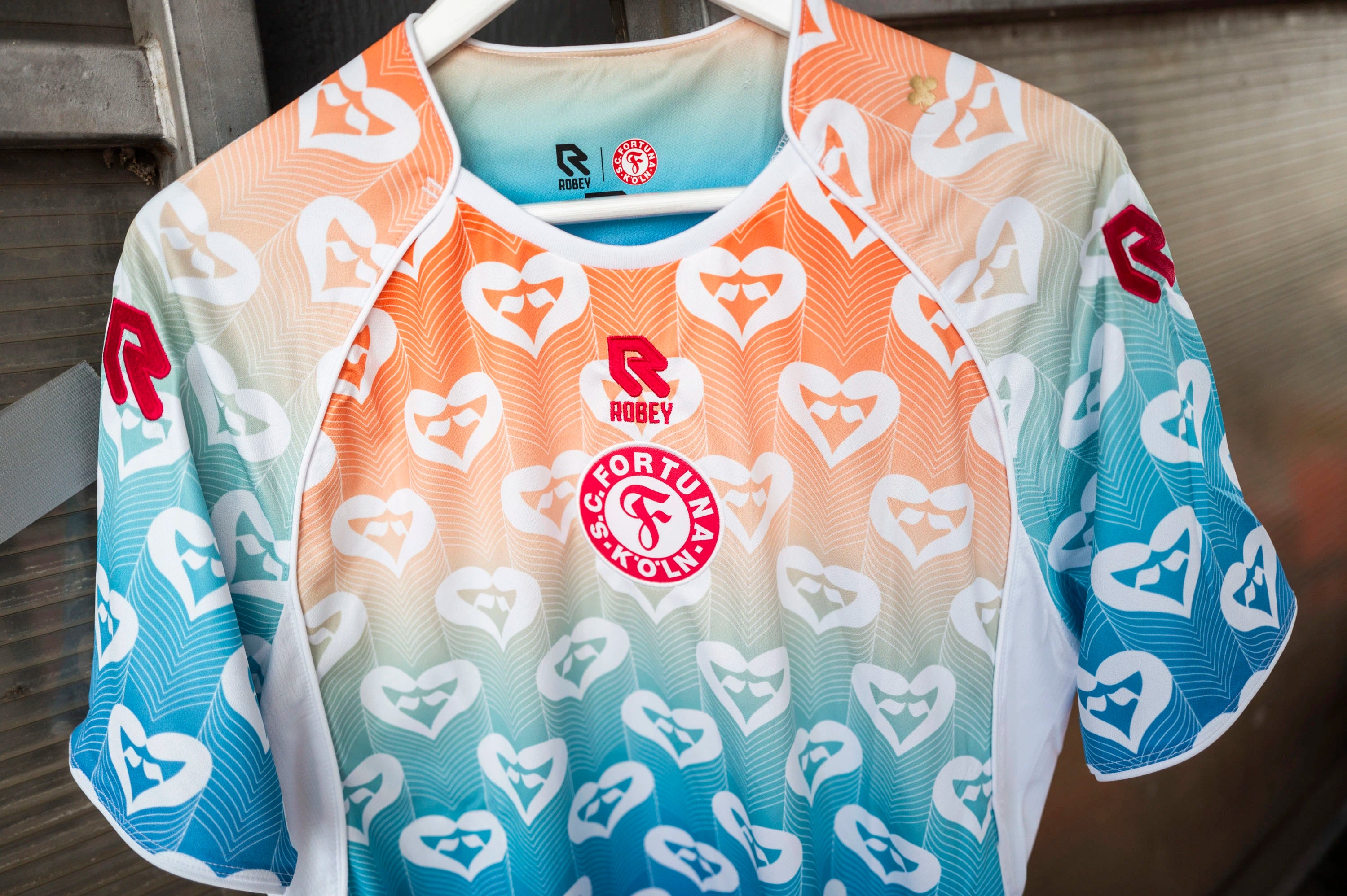 Vorderansicht des SC Fortuna Köln
Pride-Trikots, aufgenommen auf einem
Kleiderbügel. Das Design zeigt einen
sanften Farbverlauf von Orange zu Blau,
verziert mit weißen Herzsymbolen. Auf
der Brust prangen das rote Vereinslogo
und das rote Robey-Logo.