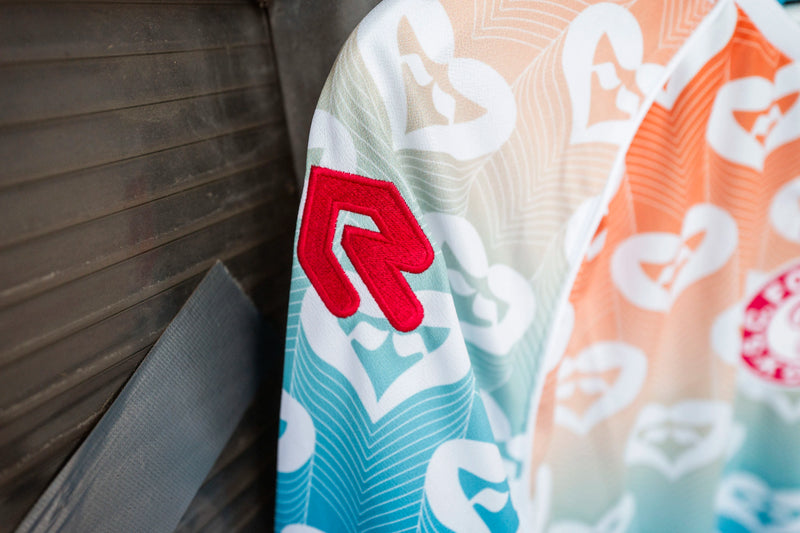 Detailaufnahme des linken Ärmels des Pride-Trikots mit dem roten Robey-Logo, weißen Herzsymbolen und dem fließenden Farbverlauf.