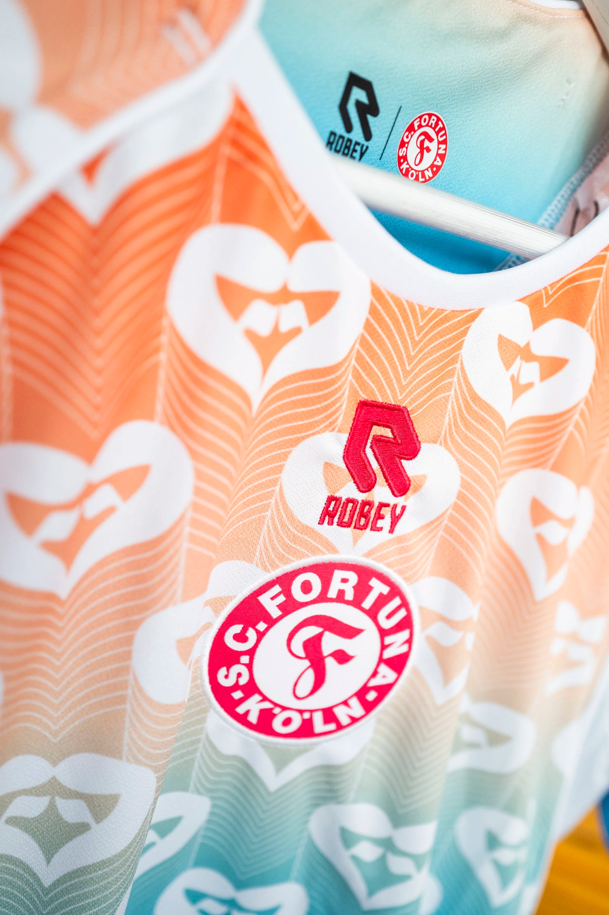 Nahaufnahme der Vorderseite des Pride-Trikots mit Fokus auf das Vereinslogo „SC Fortuna Köln“, das rote Robey-Logo und das auffällige Herzmuster im Farbverlauf.