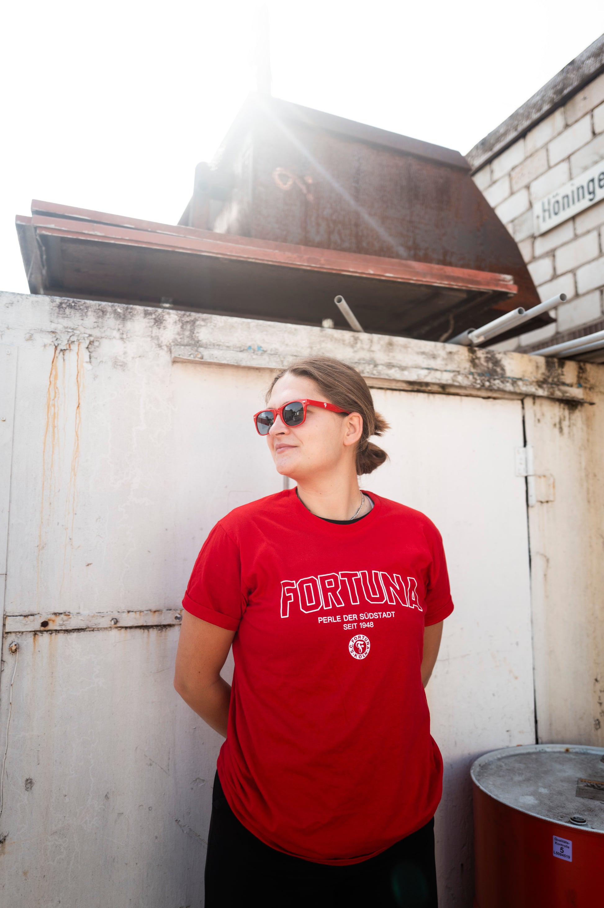 Person trägt eine rote Sonnenbrille mit Fortuna-Logo und ein rotes T-Shirt mit weißem „FORTUNA“-Schriftzug sowie Vereinslogo, lehnt sich mit verschränkten Armen an eine weiße Wand.