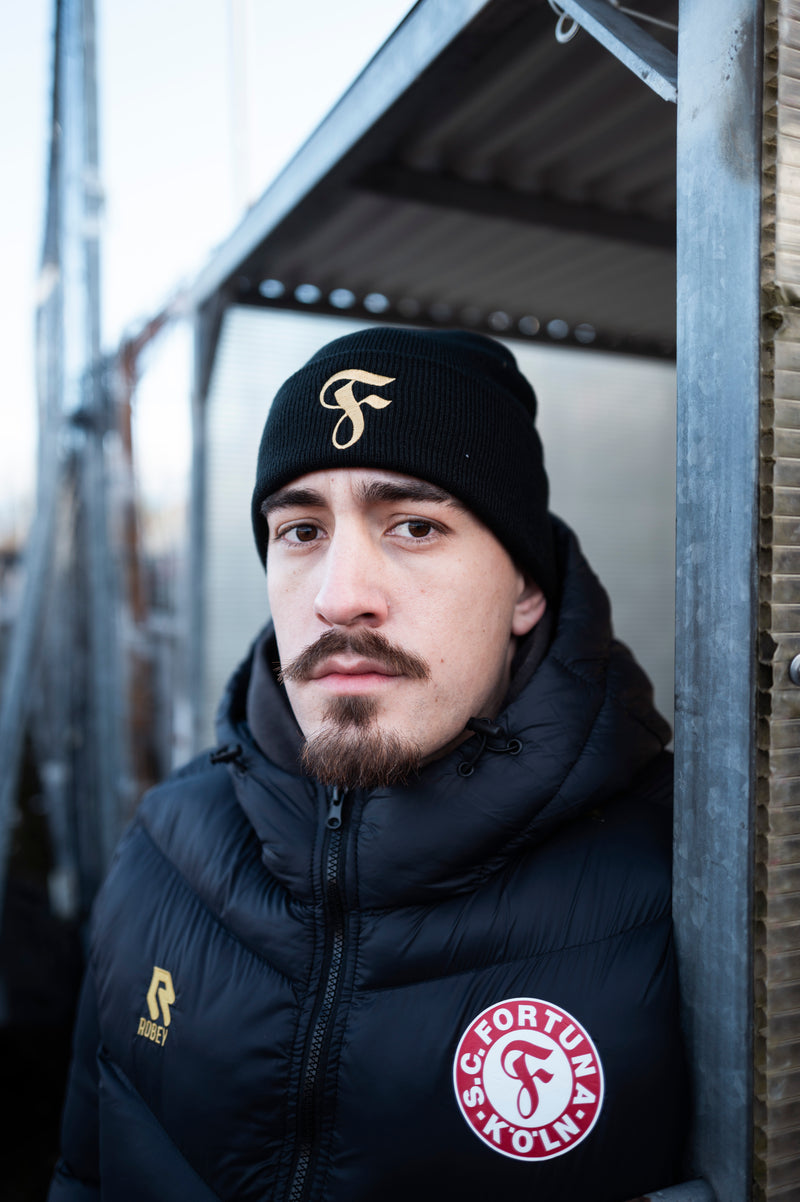 Person trägt schwarze Winterjacke
von Fortuna Köln mit rotem Vereinslogo
und goldenem Robey-Logo sowie
schwarze Mütze mit goldenem „F“-Logo.
Er steht nah an einer Metallwand und
schaut in die Kamera.