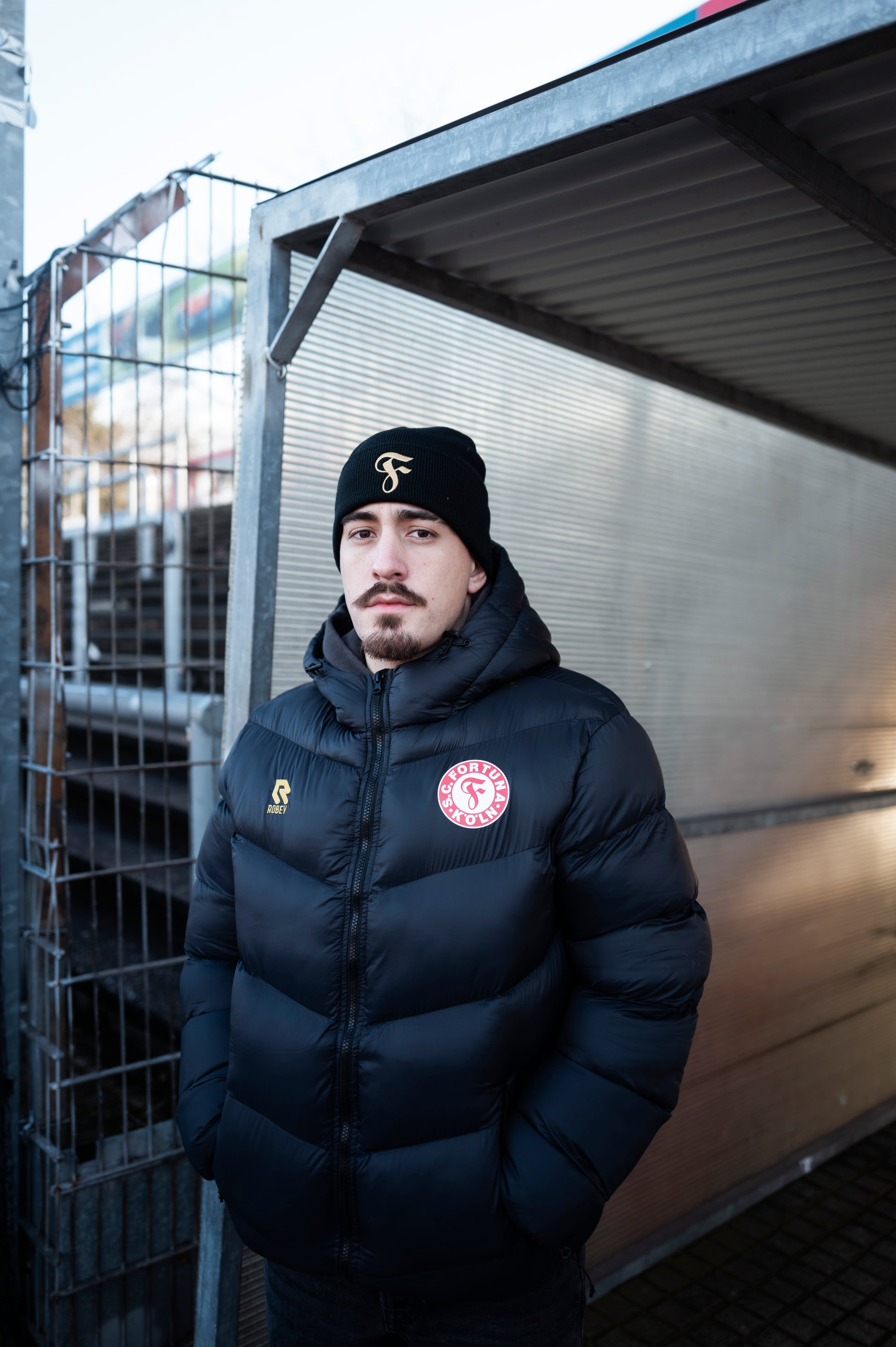 Person trägt schwarze Winterjacke von Fortuna Köln mit rotem Vereinslogo, goldenem Robey-Logo und hochgeschlossenem Kragen sowie schwarze Mütze mit goldenem „F“-Logo. Er steht unter einem überdachten Bereich und blickt nach vorn.