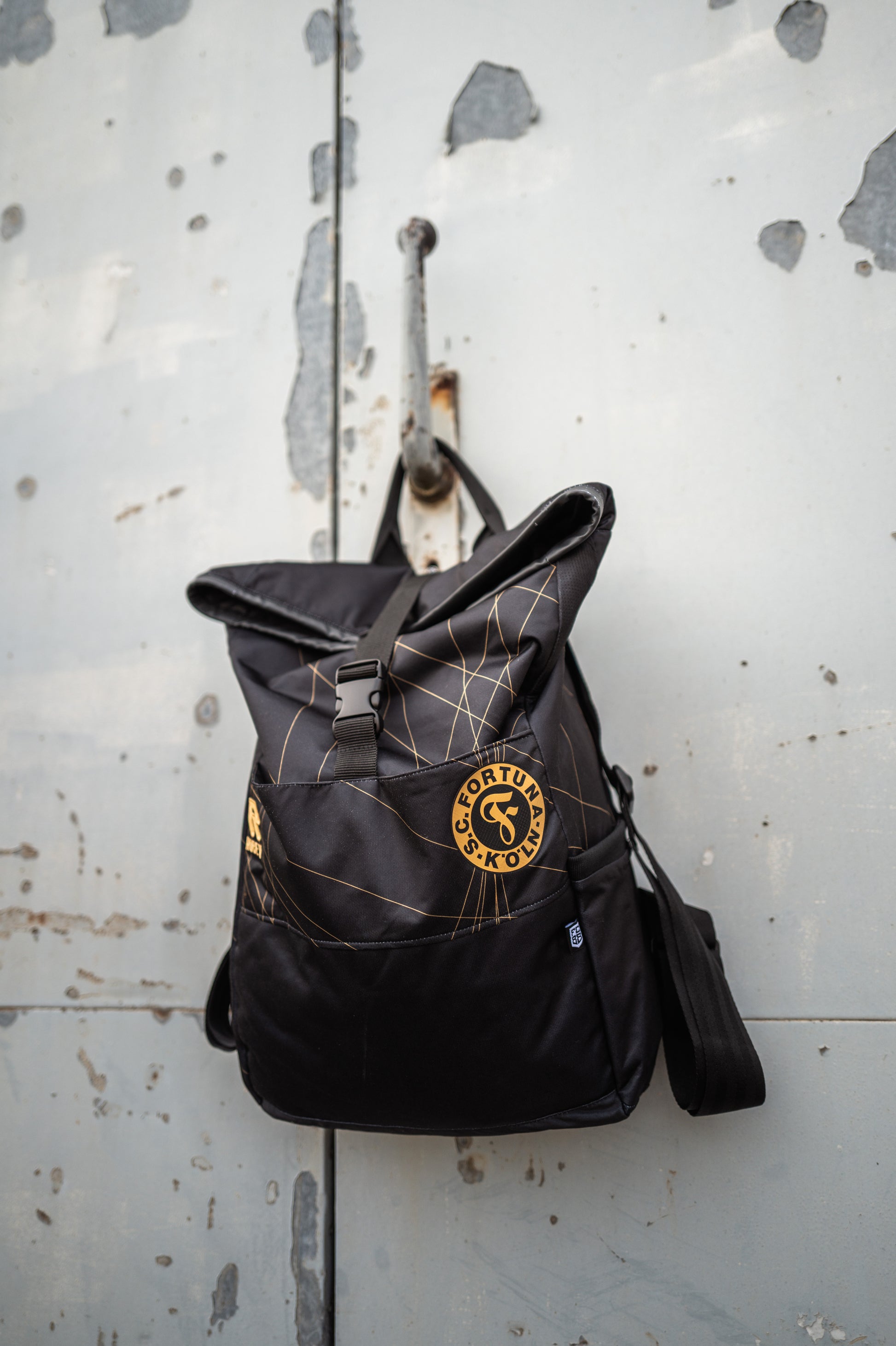 Auf dem Bild sieht man den Schwarz-goldenen Rucksack mit dem Auswärtstrikot aus der Saison 2024/2025 verarbeitet, vor einer grauen Metalltür aufgehangen.