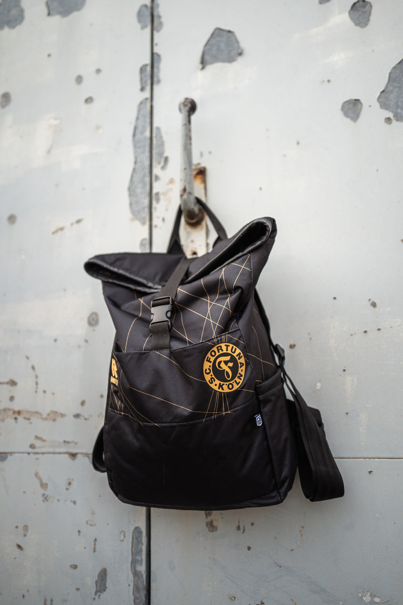 Auf dem Bild sieht man den Schwarz-goldenen Rucksack mit dem Auswärtstrikot aus der Saison 2024/2025 verarbeitet, vor einer grauen Metalltür aufgehangen.