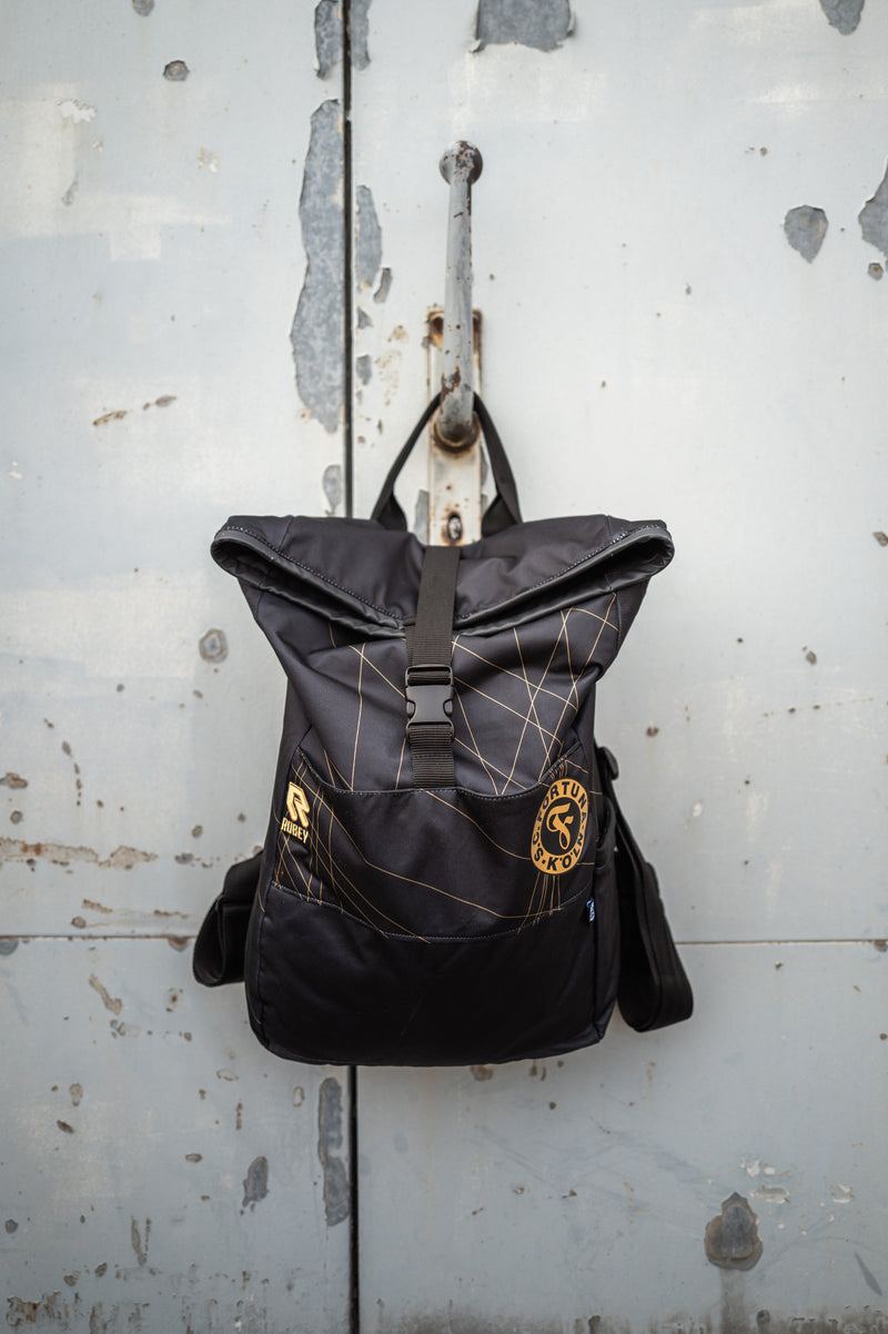 Auf dem Bild sieht man den Schwarz-goldenen Rucksack mit dem Auswärtstrikot aus der Saison 2024/2025 verarbeitet, vor einer grauen Metalltür aufgehangen.