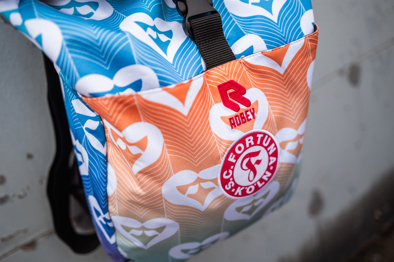 Auf dem Bild sieht man einen Rucksack, welcher aus dem Pride Shirt von Fortuna Köln verarbeitet wurde, vor einer grauen Metalltür.