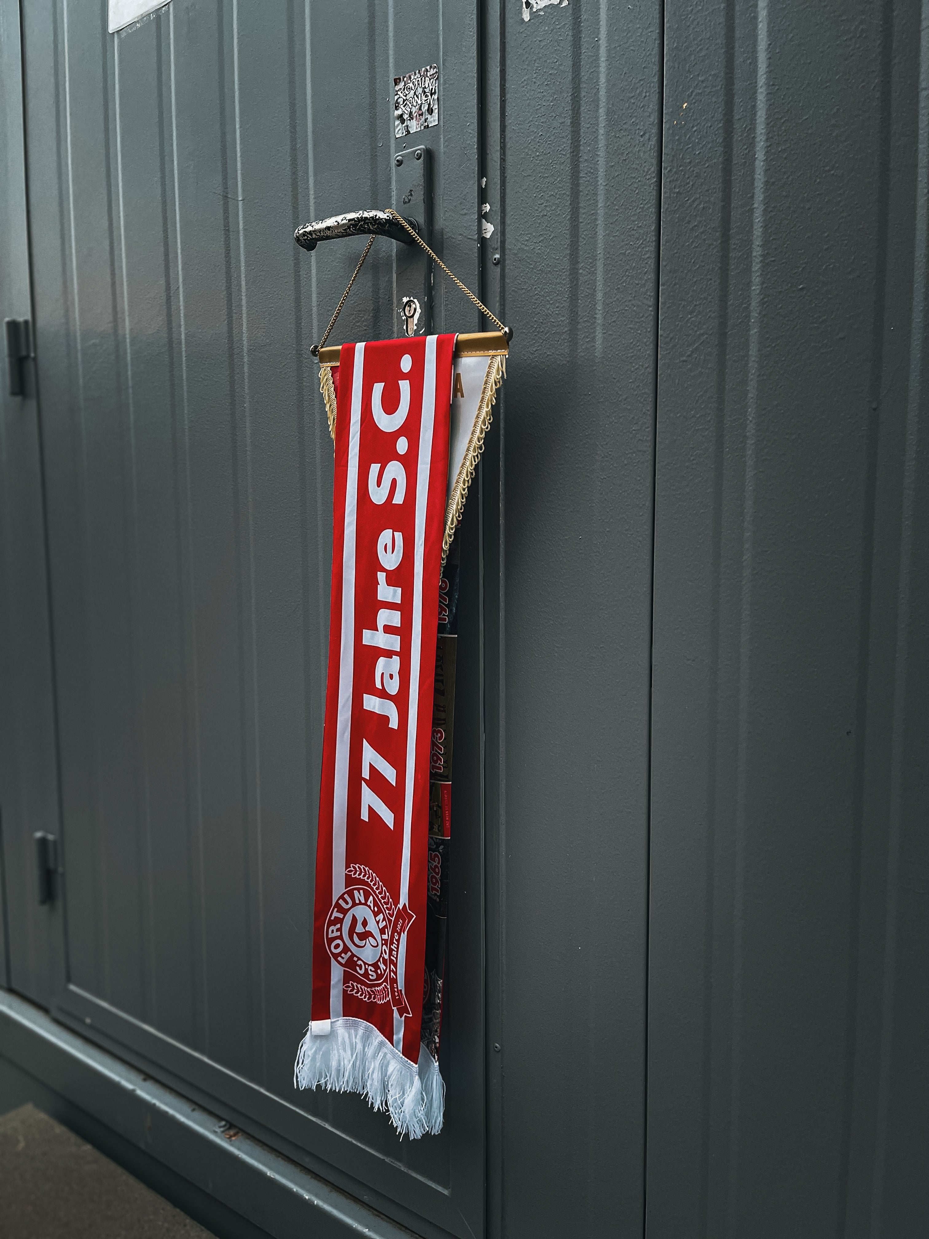 Rot-weißer Jubiläumsschal mit der Aufschrift „77 Jahre S.C.“, über einem Wimpel mit goldener Kette und weißen Fransen hängend.