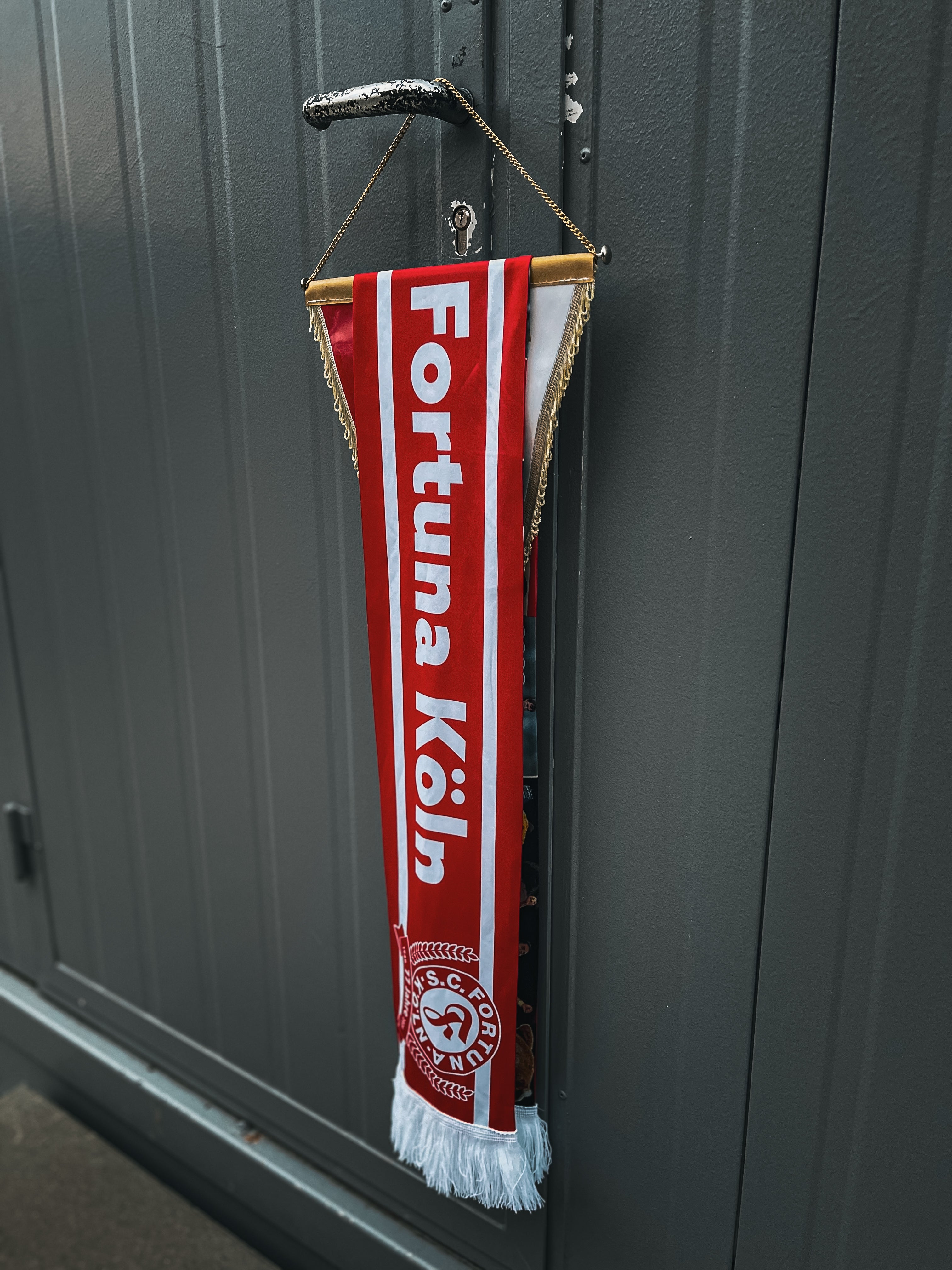 Rot-weißer Jubiläumsschal mit der Aufschrift „Fortuna Köln“, über einem Wimpel mit goldener Kette und weißen Fransen hängend.