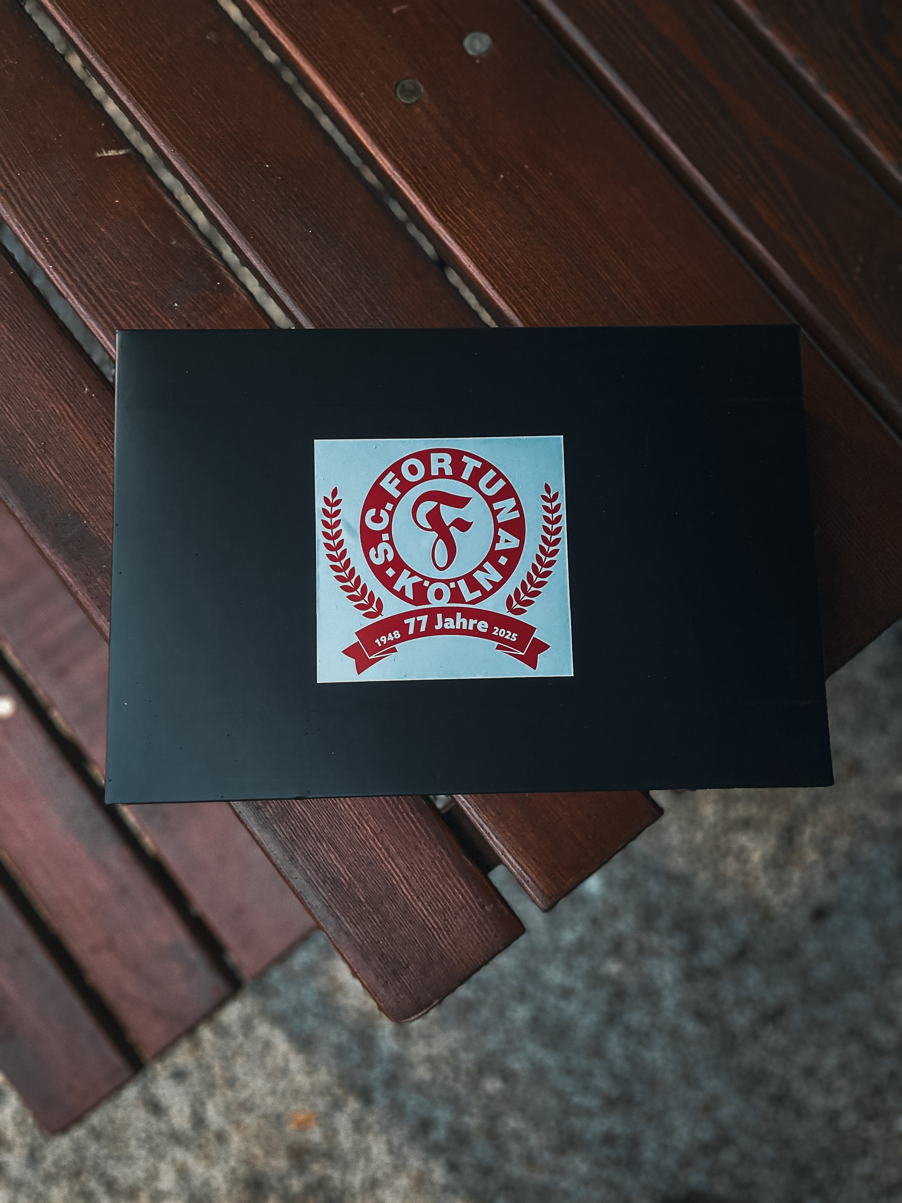 Schwarze Geschenkbox mit rotem Fortuna Köln Jubiläumslogo „77 Jahre“ auf dem Deckel.
