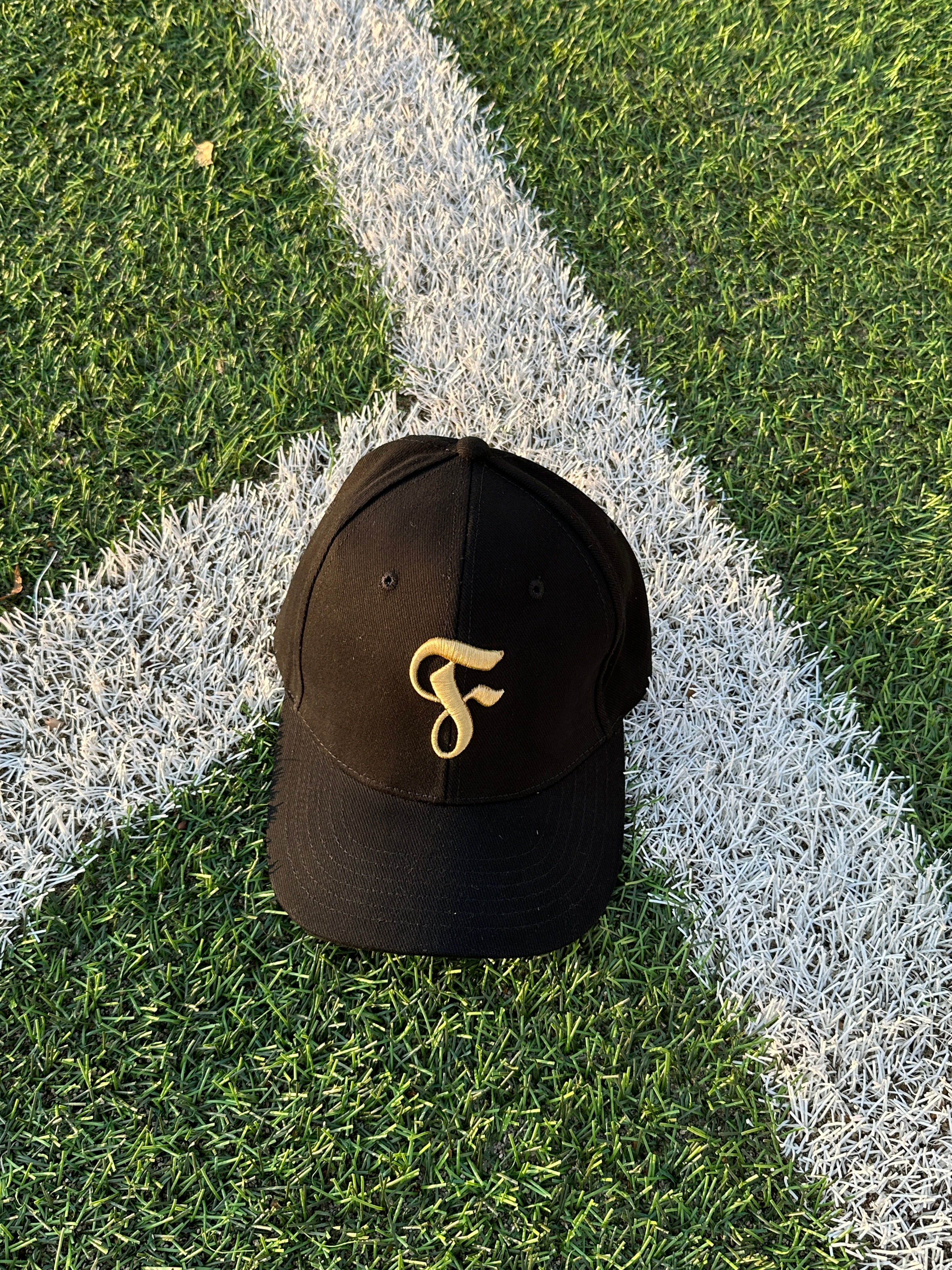 Die schwarze Cap mit goldfarbenem „F“-Logo einzeln auf grünem Kunstrasen mit weißer Spielfeldlinie.