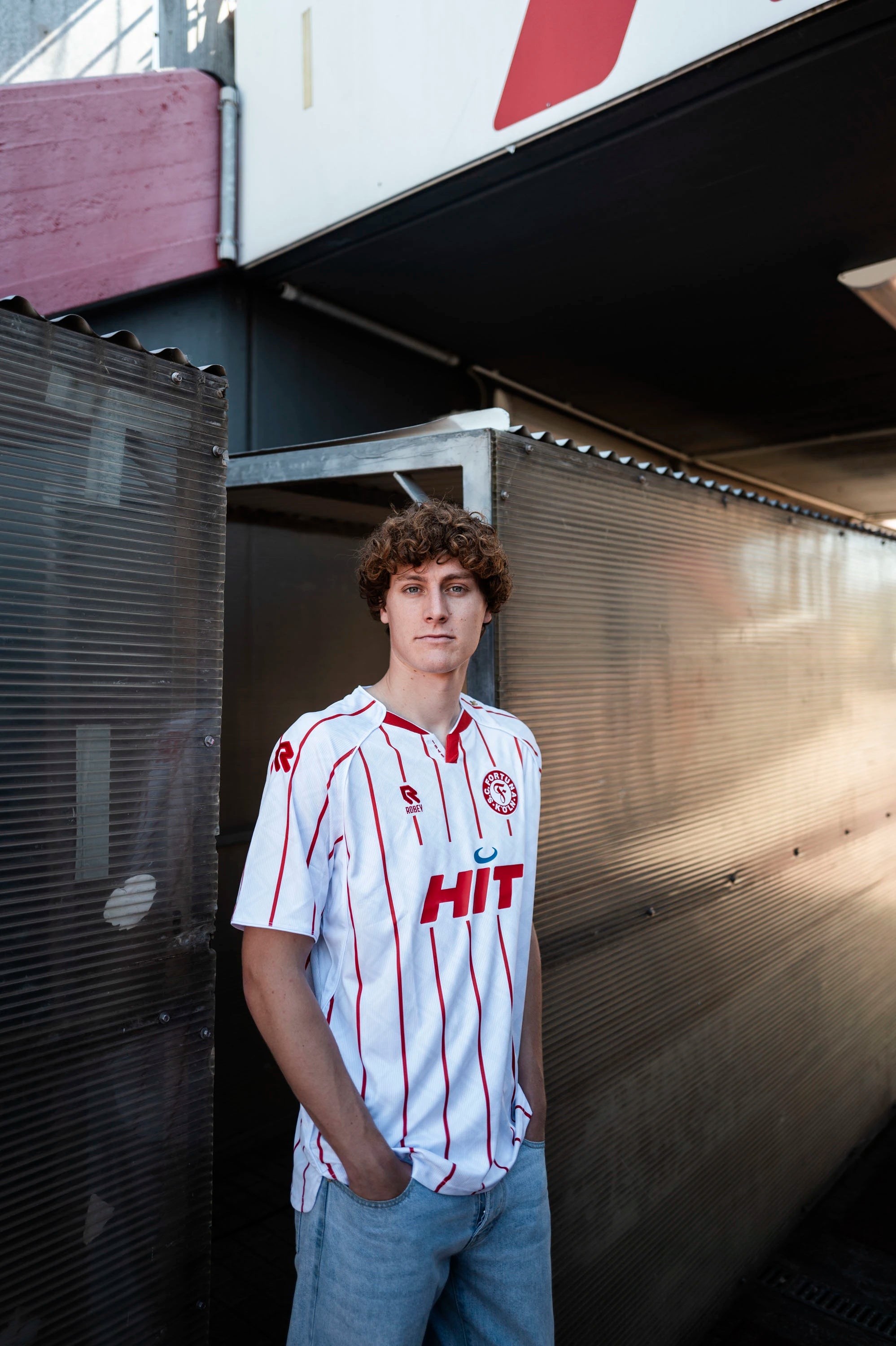 Spieler trägt das neue Heintrikot
der Saison 2025/2026. Das Trikot
ist weiß mit roten Streifen. Auf der
Brust befindet sich das rot-weiße
Fortuna-Logo sowie ein rotes
Robey-Logo, was sich ebenfalls
auf der Schulter finden lässt.
Zentral auf dem Trikot befindet
sich ein großes Sponsoren-Logo
von HIT.