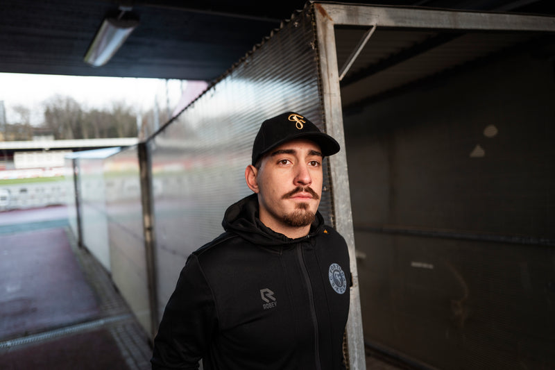 Eine Person trägt die Cap zur schwarzen Robey-Trainingsjacke mit Fortuna-Logo, Aufnahme im Stadionbereich unter einem überdachten Gang.