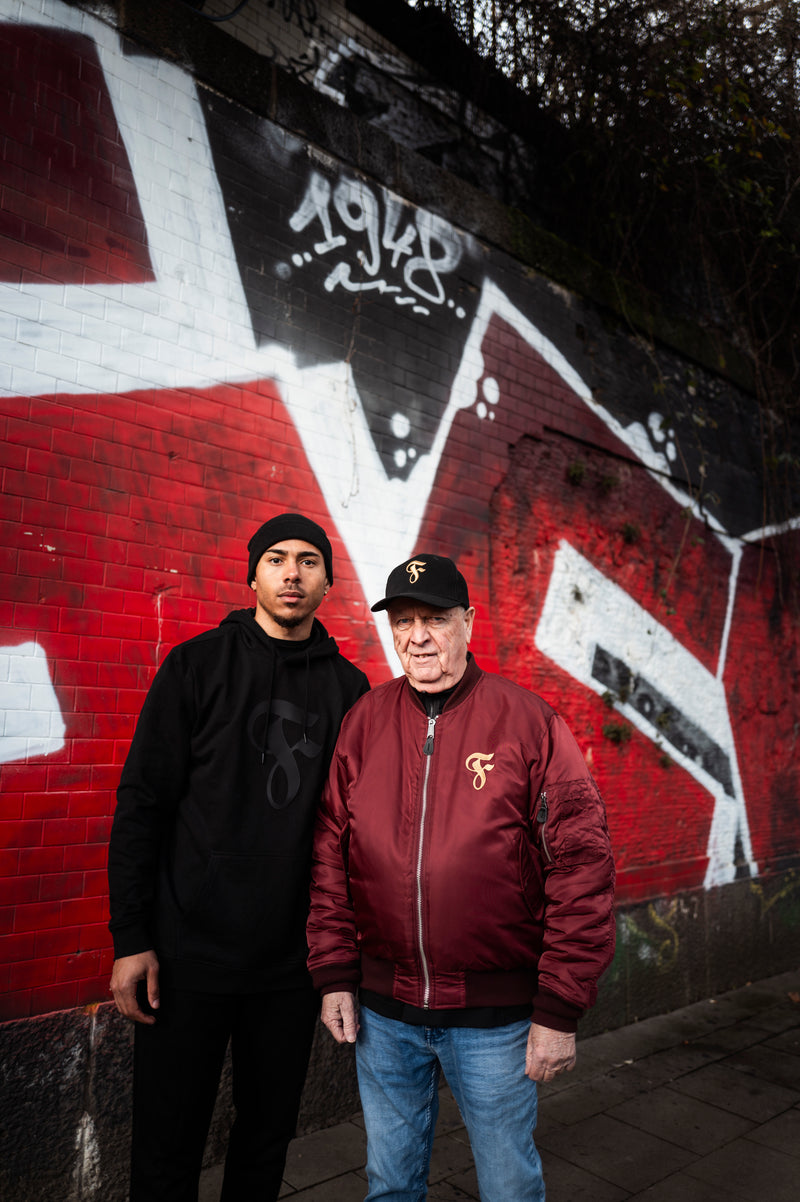 Eine Person steht neben einem Fortuna-Spieler, trägt dabei ebenfalls die schwarze Cap, Bomberjacke, schwarzes Oberteil und Jeans. Hintergrund: rote und weiße Graffiti-Mauer mit „1948“-Schriftzug.