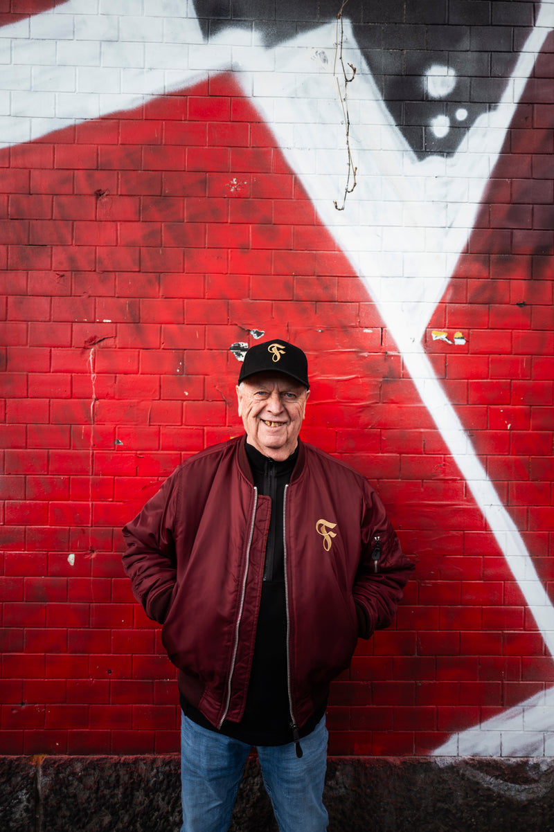Eine Person trägt die schwarze Cap mit goldfarbenem „F“-Logo, dazu eine bordeauxrote Fortuna-Bomberjacke mit passendem Logo, schwarzes Oberteil und blaue Jeans. Hintergrund: rote Graffiti-Mauer.
