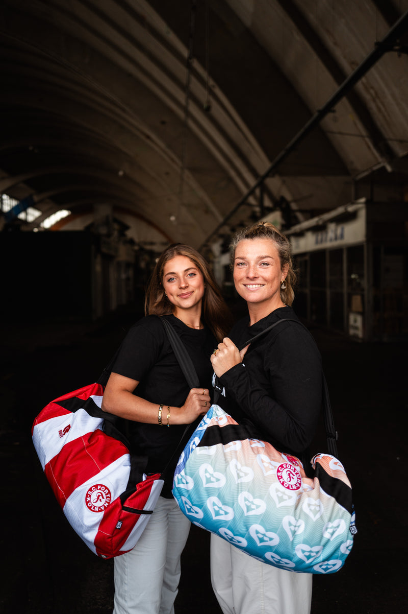 Auf dem Bild sieht man zwei Damen. Die Linke Dame trägt die Wochenender Tasche mit dem Heimtrikot der Saison 2024/2025. Die rechte Dame trägt die Wochenender Tasche, welches aus dem Pride Shirt.