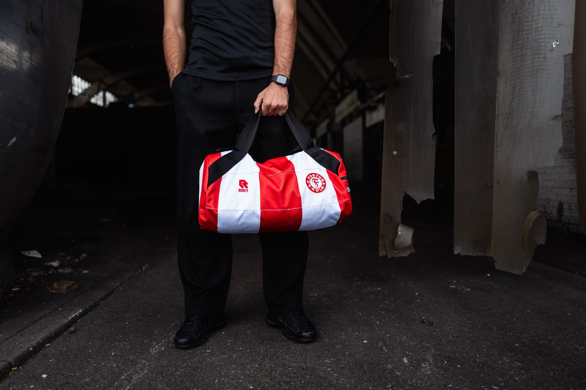 Man sieht einen Mann der die Tasche aus dem alten Heimtrikot trägt. Die Tasche ist Rot-Weiß gestreift und trägt das Robey Logo links und das Fortuna Wappen rechts auf dem Bild.