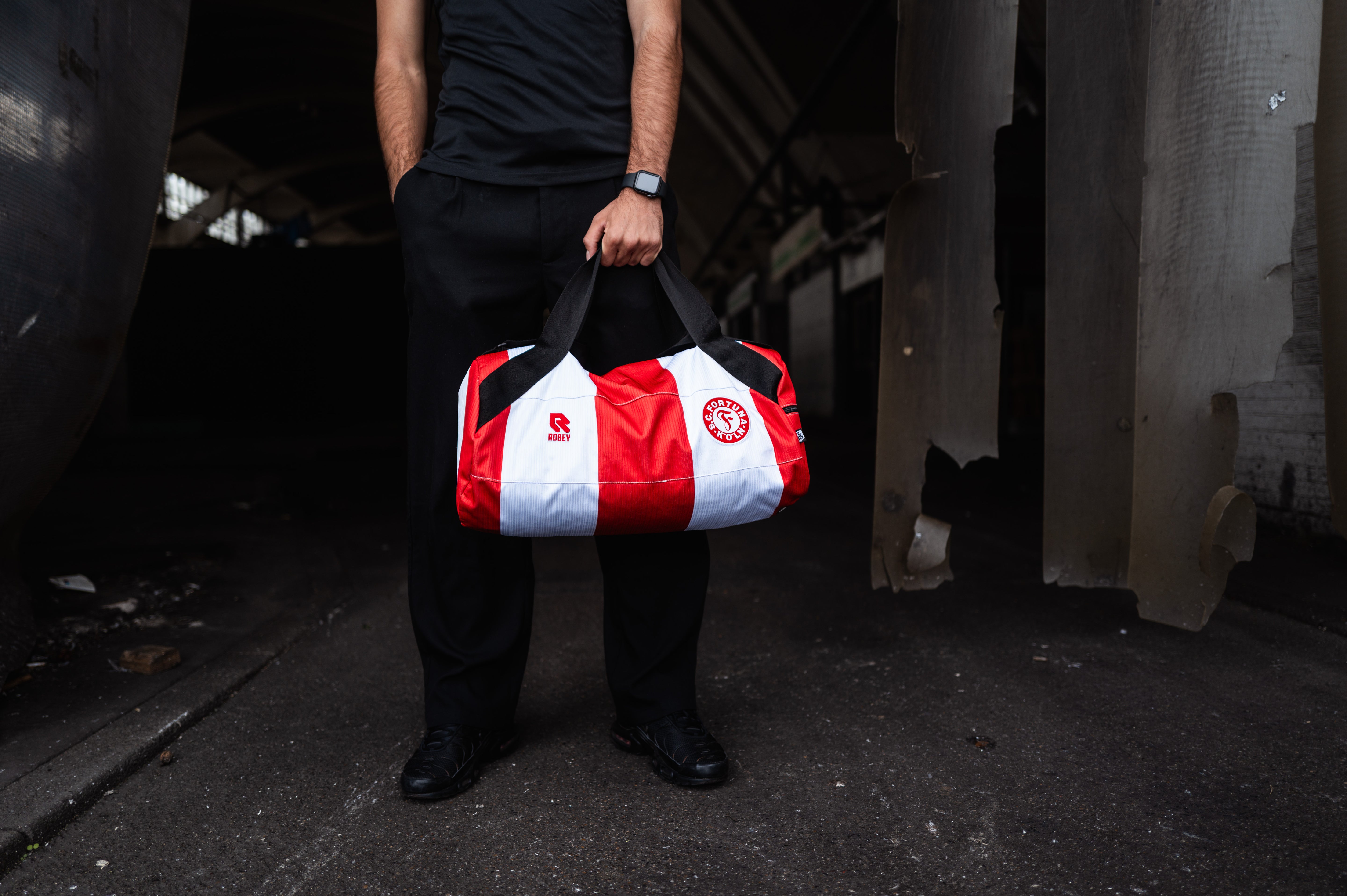Man sieht einen Mann der die Tasche aus dem alten Heimtrikot trägt. Die Tasche ist Rot-Weiß gestreift und trägt das Robey Logo links und das Fortuna Wappen rechts auf dem Bild.