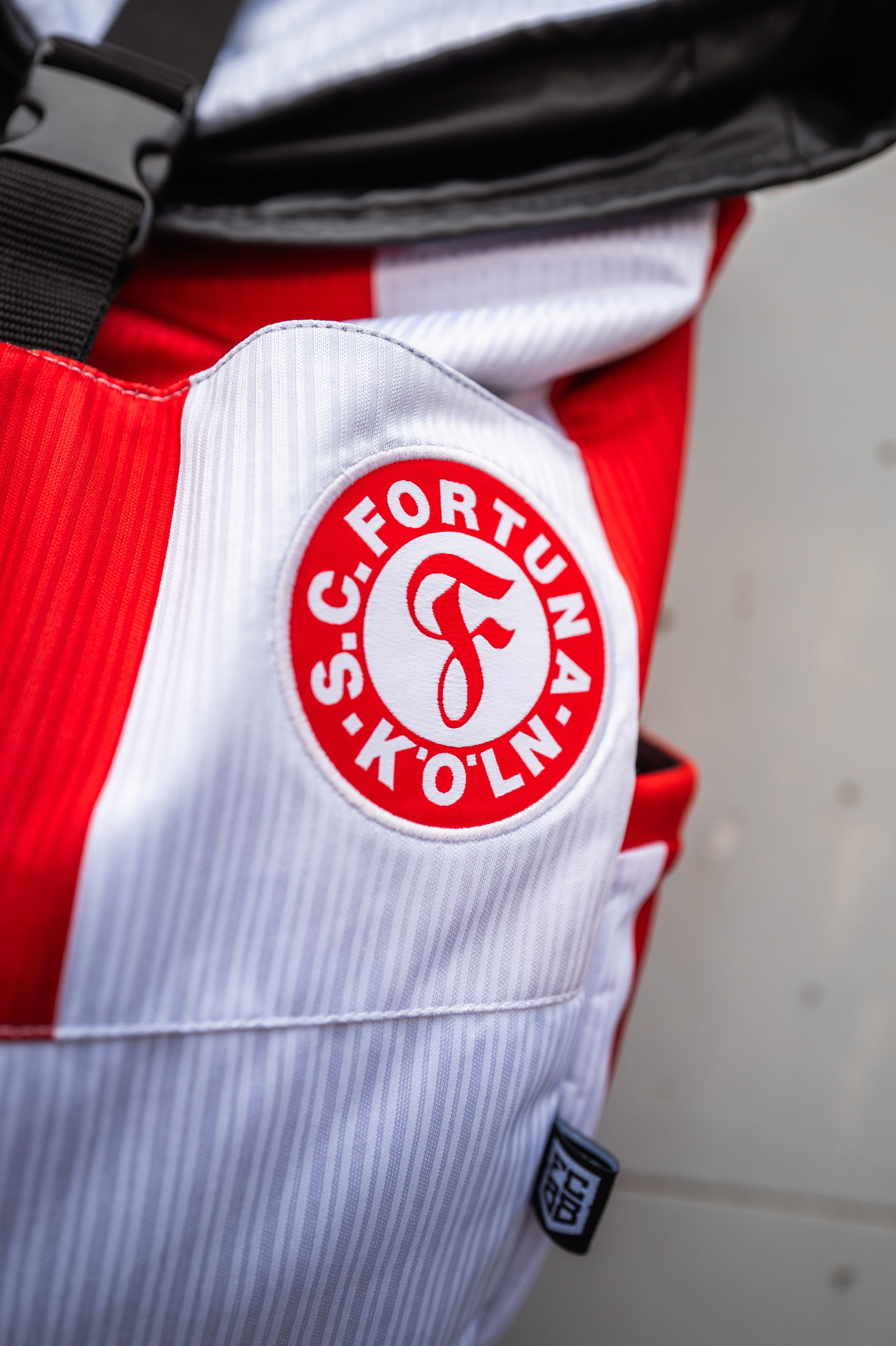 Auf dem Bild sieht man das Rot-Weiße Fortuna Köln Logo auf einem Rucksack. Der Rucksack ist eine Nahaufnahme, welcher Rot-Weiße Streifen hat.
