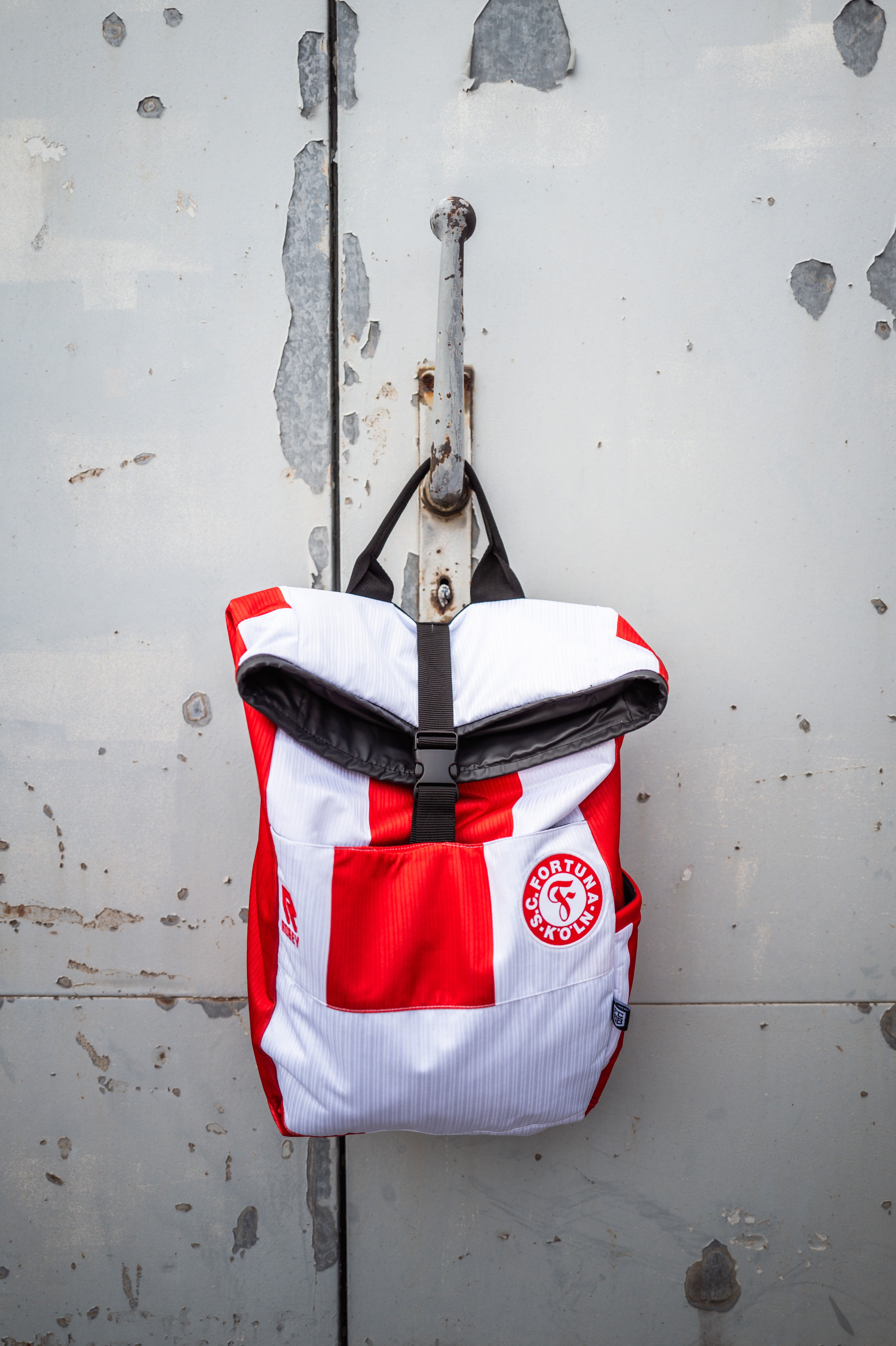 Auf dem Bild ist ein Rot-Weiß gestreifter Rucksack vor einer grauen Metalltür. Auf dem Rucksack ist das Heimtrikot der Saison 2024/2025 verarbeitet. 