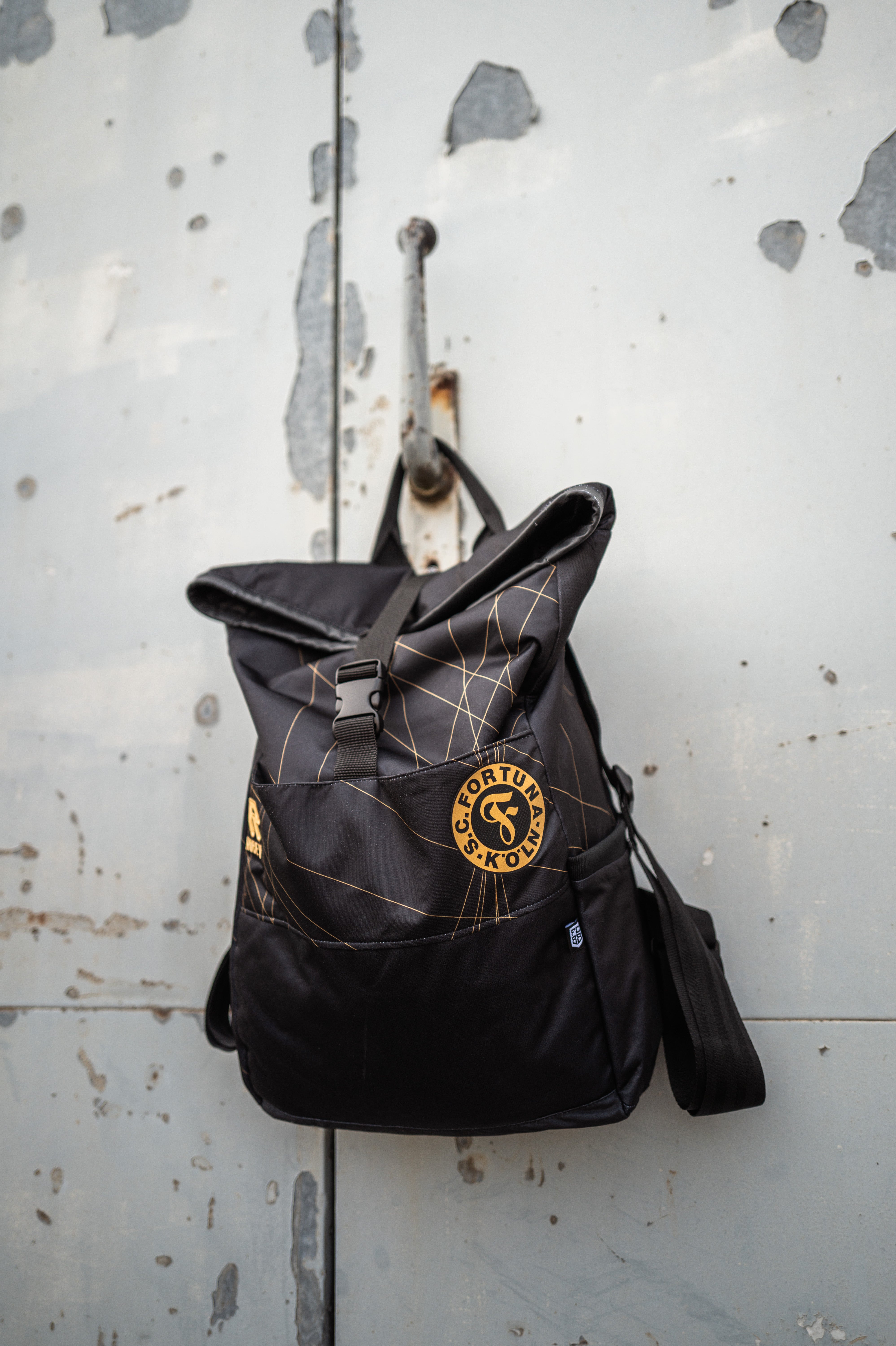 Auf dem Bild sieht man den Schwarz-goldenen Rucksack mit dem Auswärtstrikot aus der Saison 2024/2025 verarbeitet, vor einer grauen Metalltür aufgehangen.