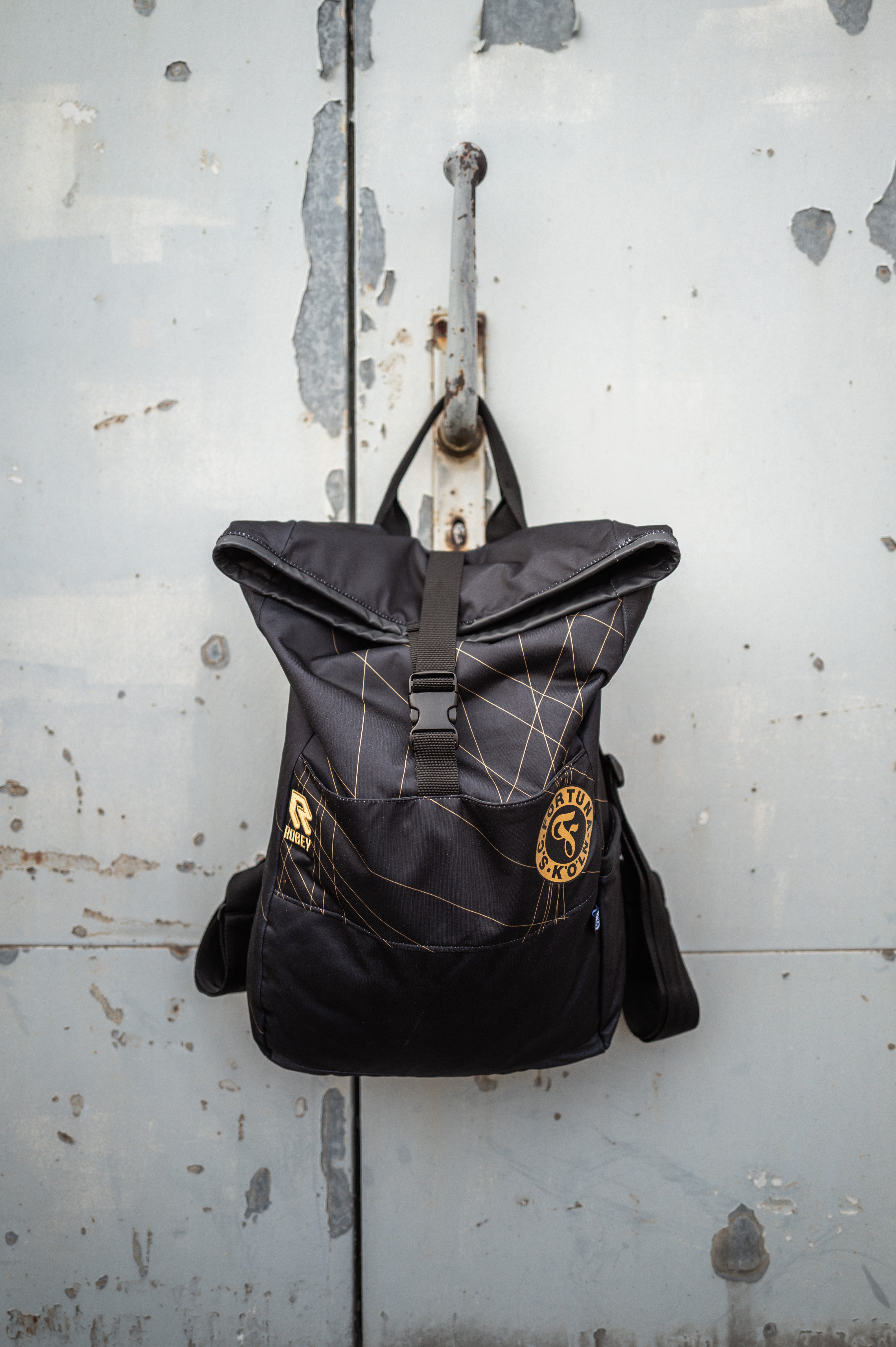 Auf dem Bild sieht man den Schwarz-goldenen Rucksack mit dem Auswärtstrikot aus der Saison 2024/2025 verarbeitet, vor einer grauen Metalltür aufgehangen. 