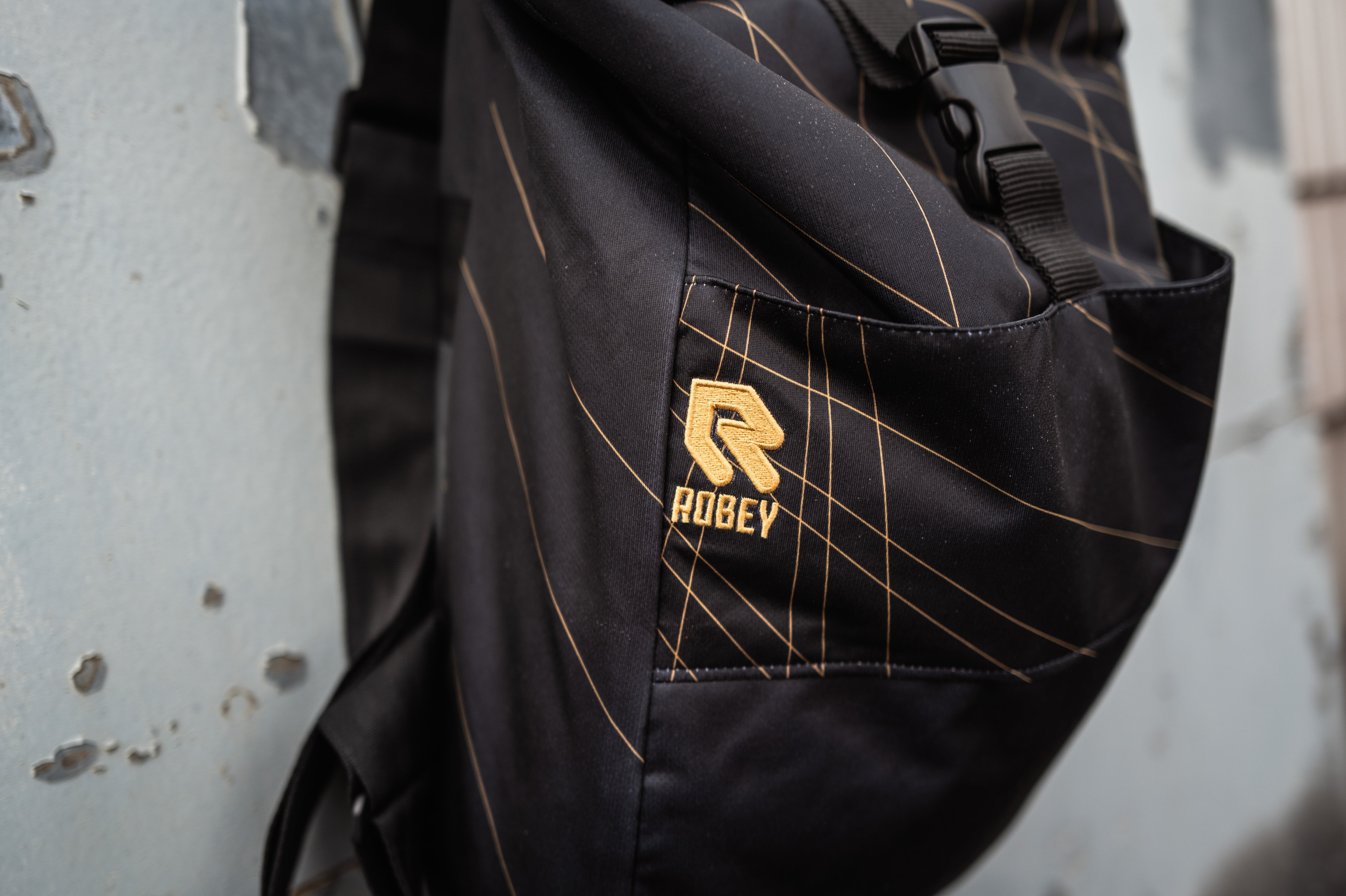 Auf dem Bild sieht man den Schwarz-goldenen Rucksack mit dem Auswärtstrikot aus der Saison 2024/2025 verarbeitet, vor einer grauen Metalltür aufgehangen.