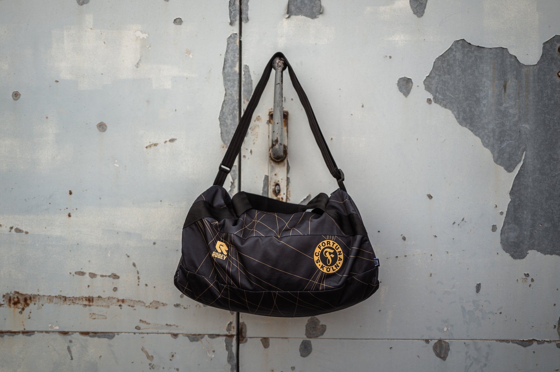 Auf diesem Bild sieht man die Schwarze Wochenender Tasche an einer Grauen Metalltür am Hacken hängen. 