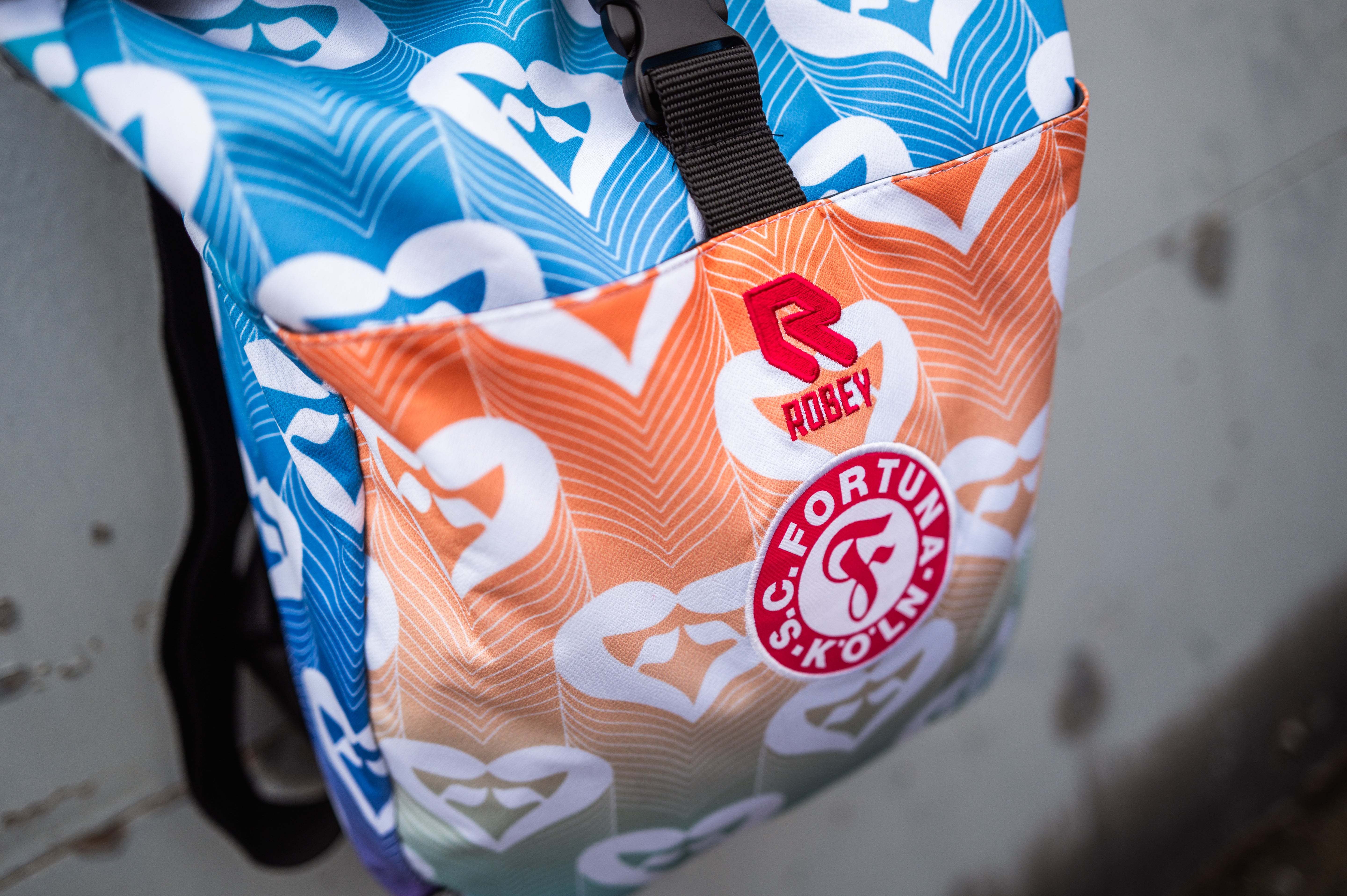Auf dem Bild sieht man einen Rucksack, welcher aus dem Pride Shirt von Fortuna Köln verarbeitet wurde, vor einer grauen Metalltür.