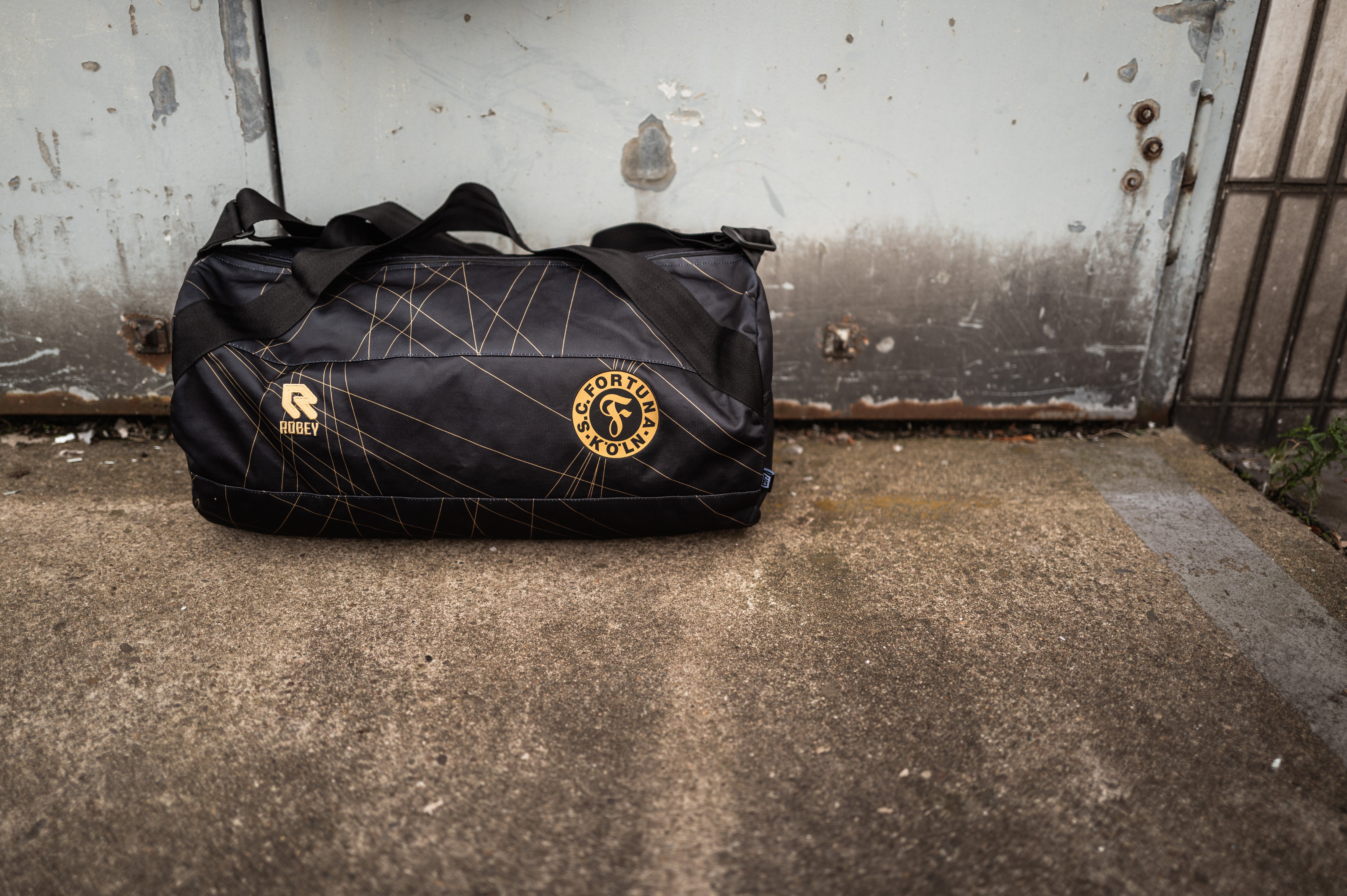 Die Wochenender Tasche aus dem Auswärtstrikot 2024/2025. Das Trikot ist hauptsächlich schwarz und hat goldene Akzente. Außerdem ist das Wappen aus Schwarz und Gold. Im Hintergrund sieht man eine graue Metalltür.