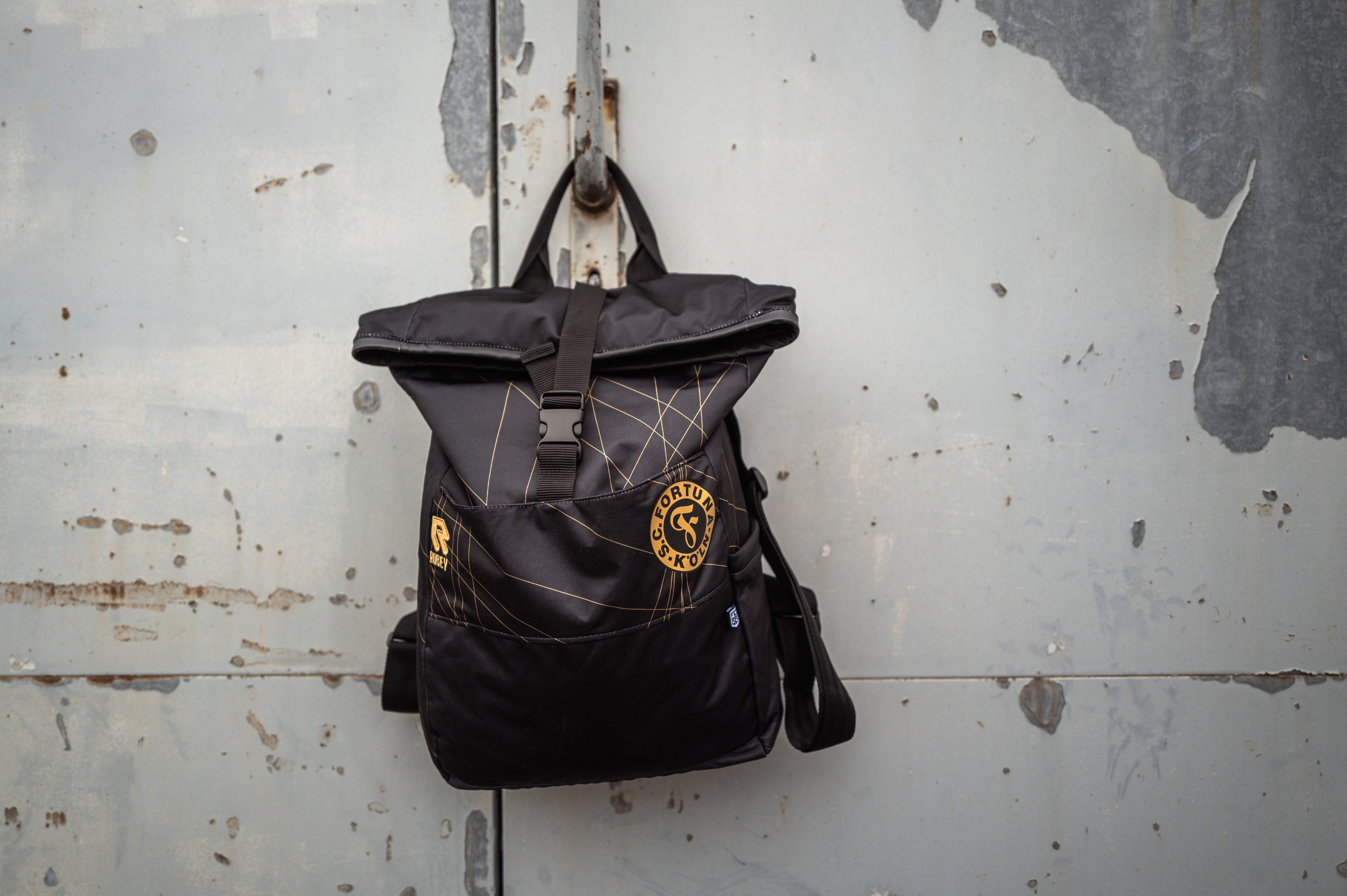 Auf dem Bild sieht man den Schwarz-goldenen Rucksack mit dem Auswärtstrikot aus der Saison 2024/2025 verarbeitet, vor einer grauen Metalltür aufgehangen.