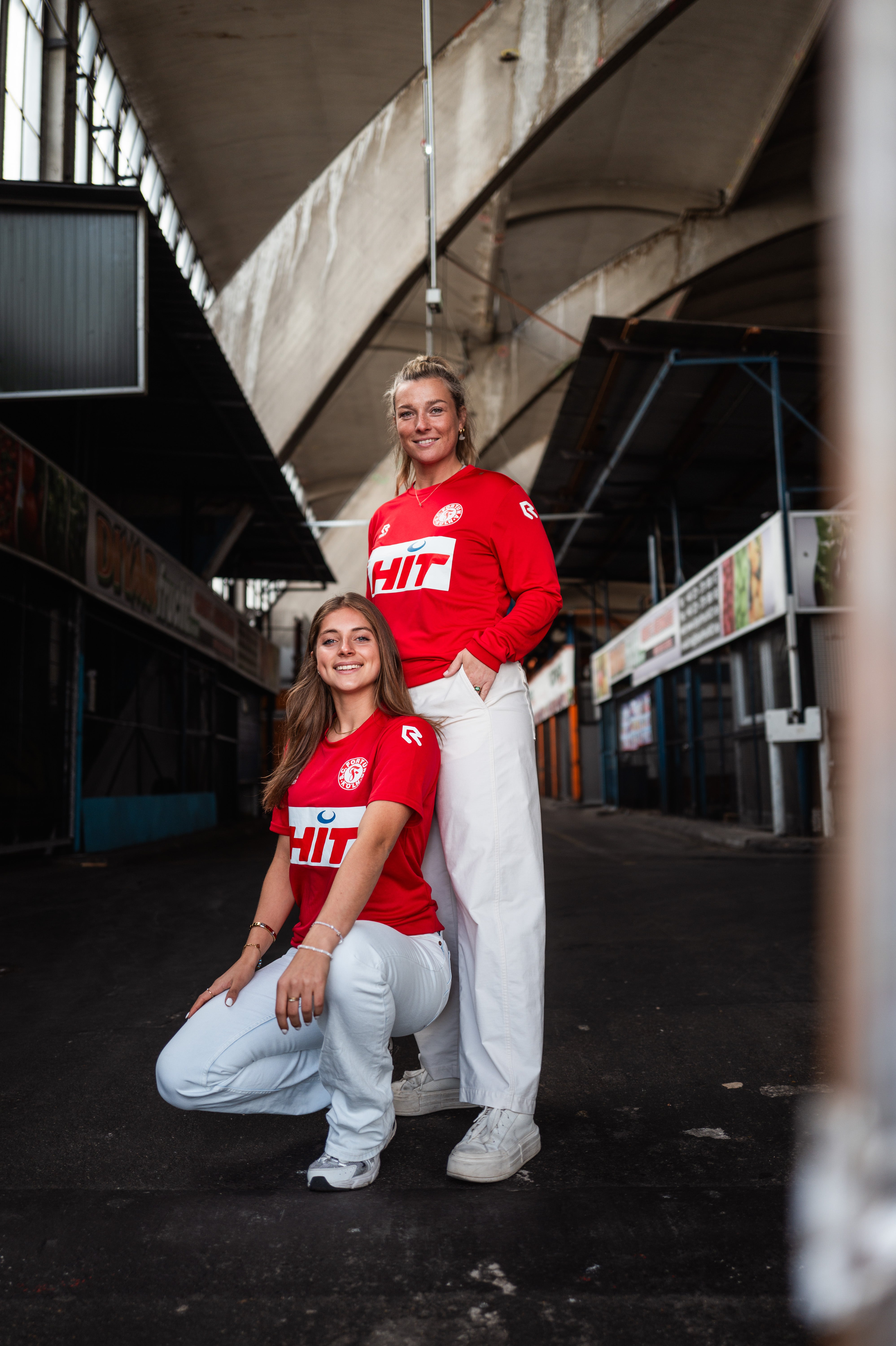 Eine Dame trägt das langarm-Trainingsshirt von Fortuna Köln. Vor ihr hockt eine Dame die das kurzarm Trainingshirt trägt.