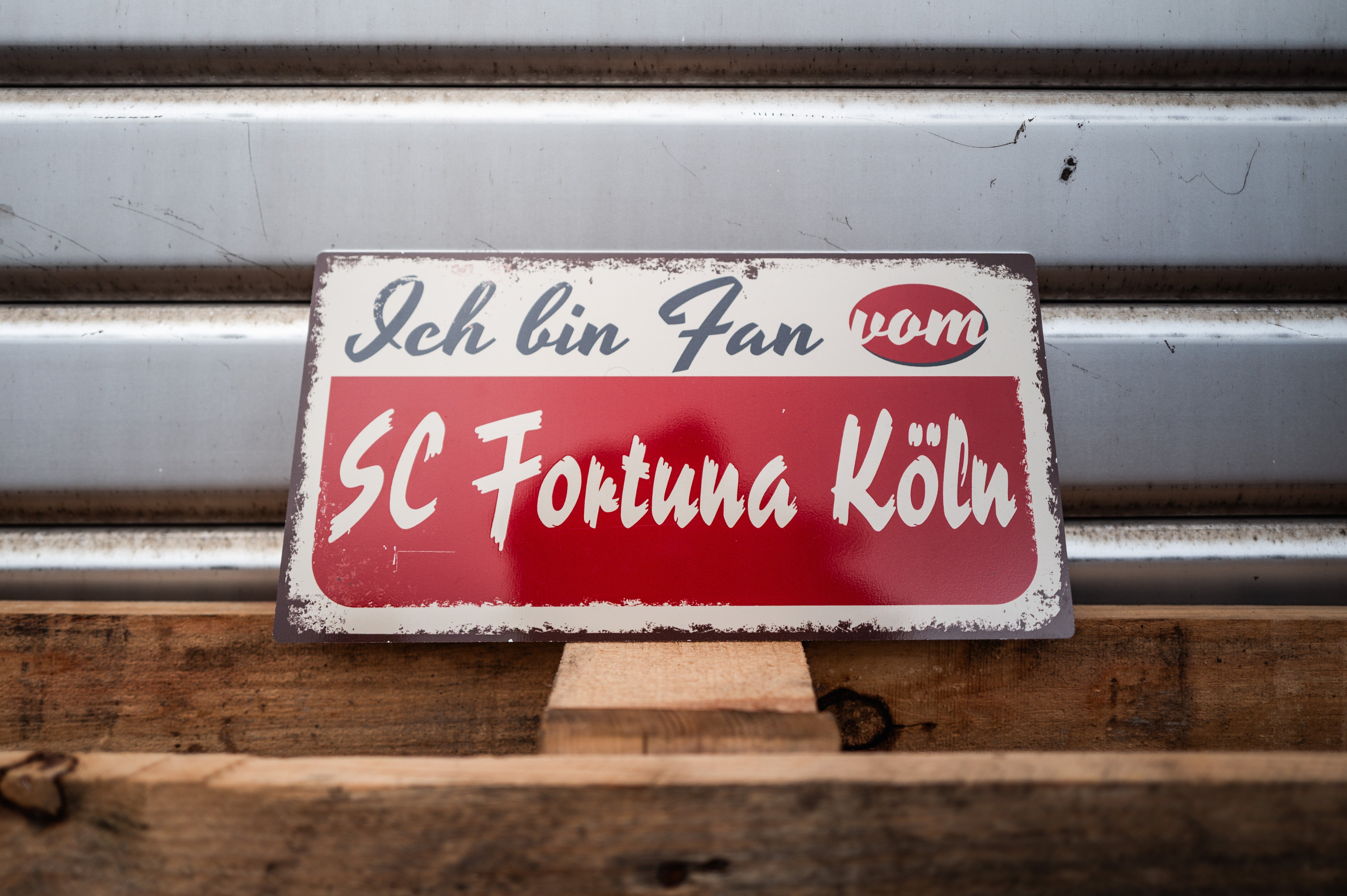 Ein Metallschild mit der Aufschrift "Ich bin Fan von SC Fortuna Köln" auf einer Holzpalette die vor einem Garagentor 